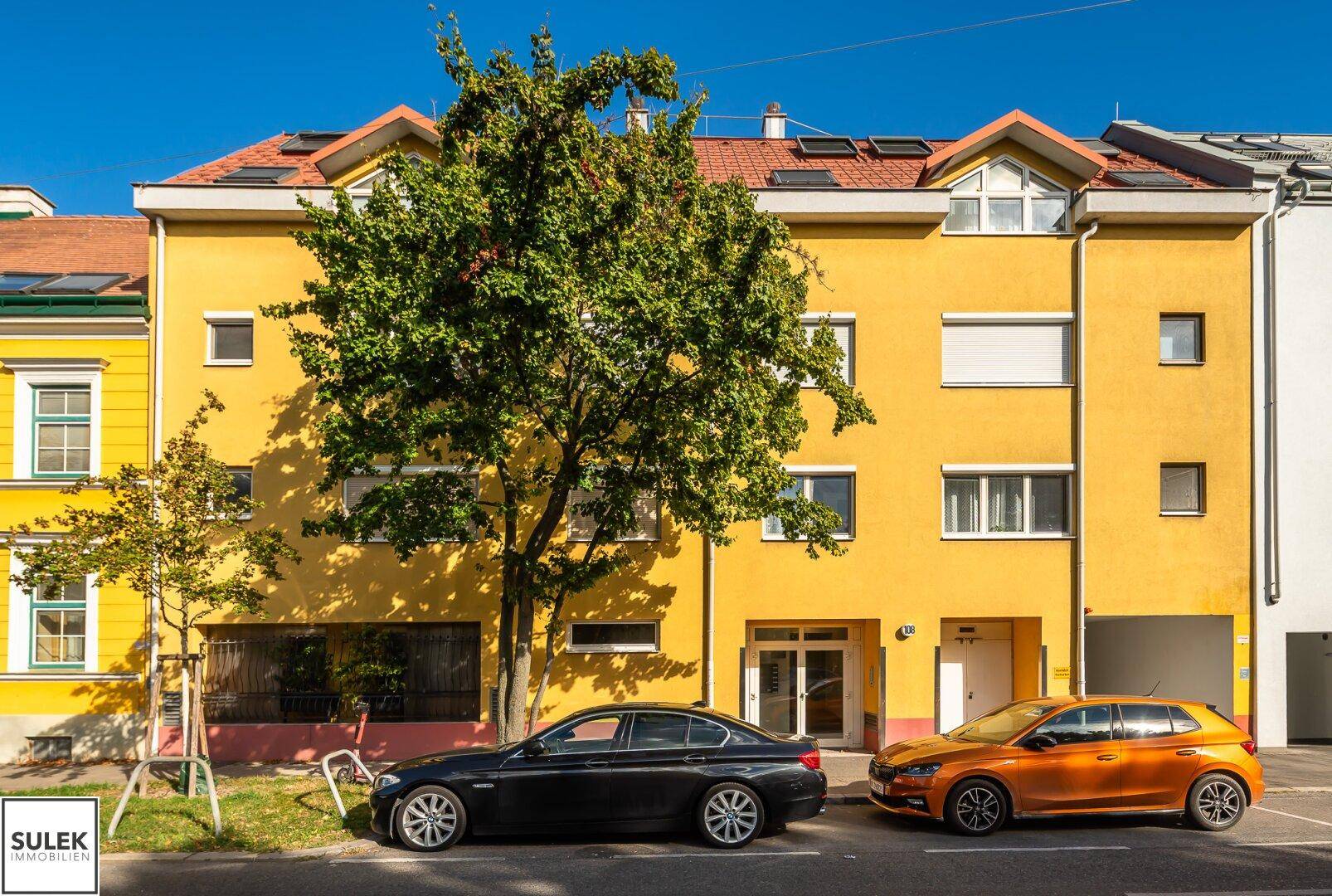 Gelbe Gebäudefassade mit mehreren Fenstern und geparkten Autos davor.