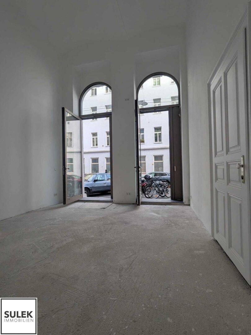 Heller, leerer Raum mit hohen Decken und zwei großen Rundbogenfenstern zur Straße hin.