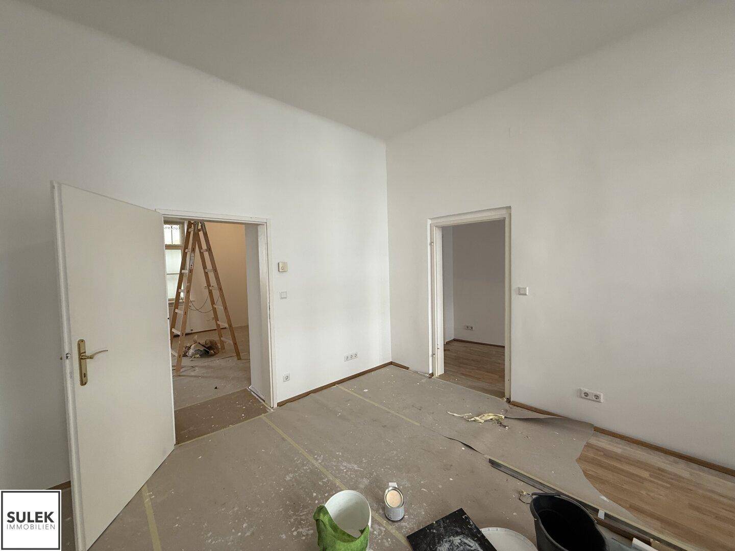 Renovierungsbedürftiger Innenraum mit weißen Wänden und zwei Türöffnungen zu angrenzenden Zimmern.
