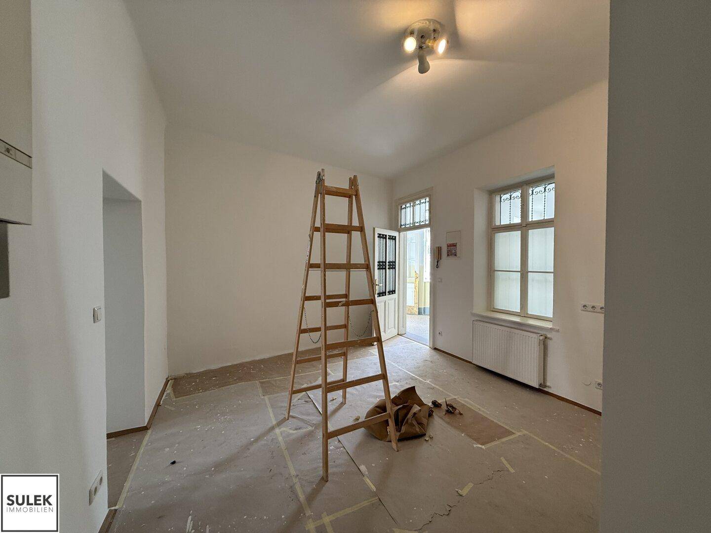Renovierungsbedürftiger Raum mit Leiter, weißen Wänden und einer Tür mit Glaseinsätzen, die nach außen führt.