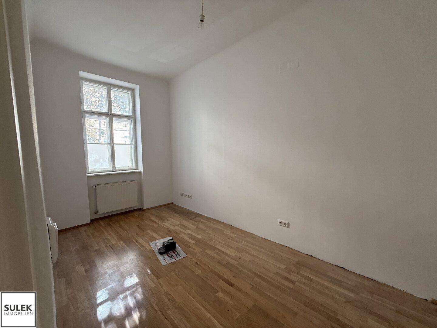 Helles Zimmer mit Holzboden und einem großen Fenster, das viel Tageslicht hereinlässt.