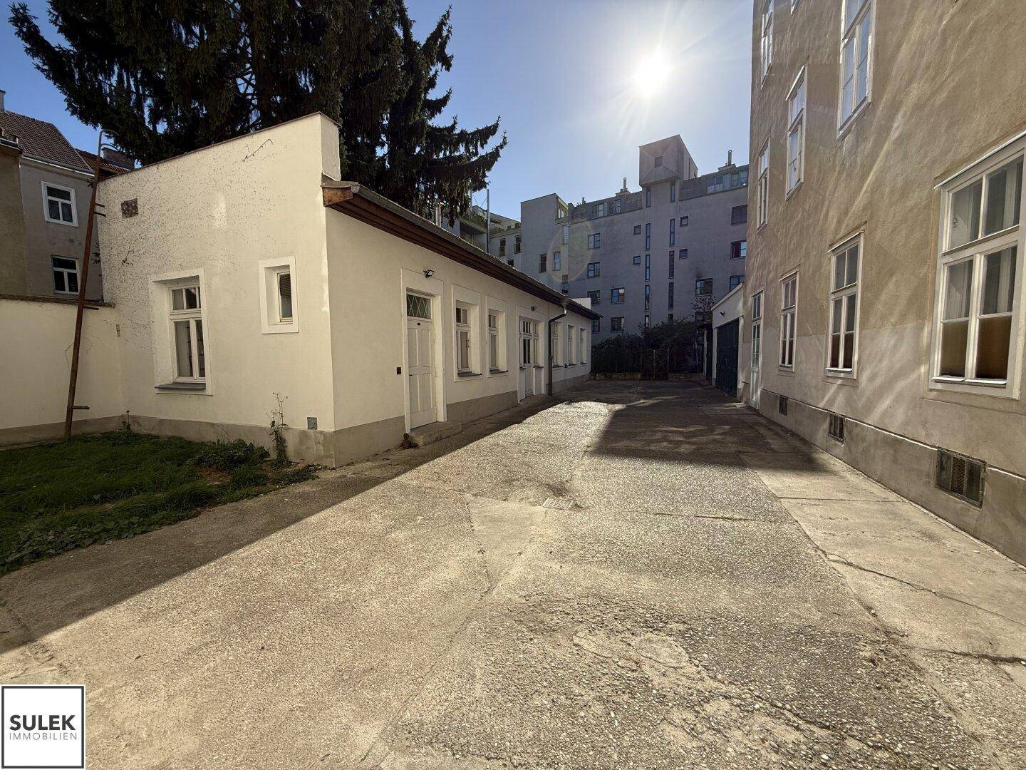 Kleines Gebäude in einem Hinterhof mit Betonfläche und weiteren Gebäuden im Hintergrund unter strahlendem Sonnenschein.