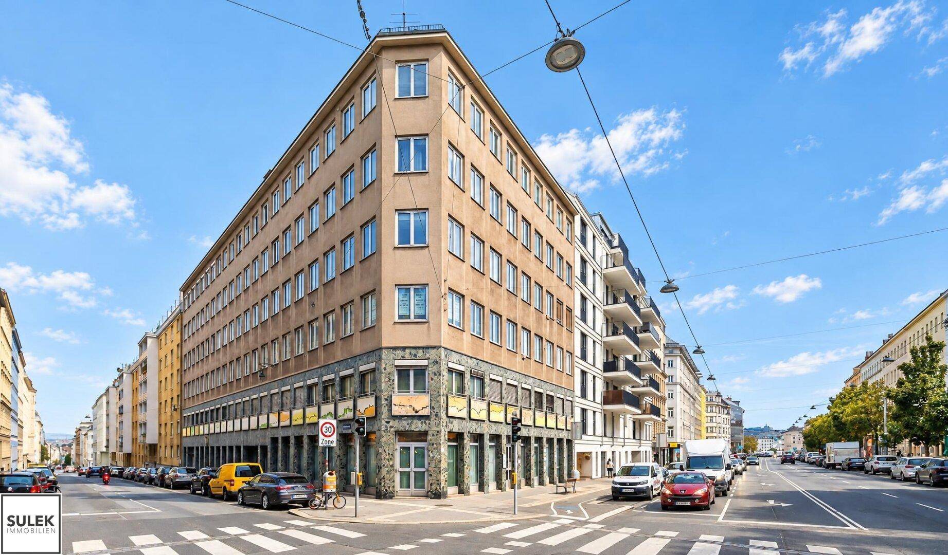 Eckgebäude in der Stadt mit mehreren Etagen, Balkonen und Geschäften im Erdgeschoss.