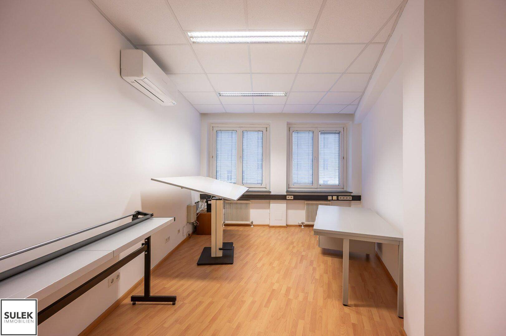 Geräumiges Büro mit Schreibtischen, Klimaanlage und großen Fenstern mit Jalousien.