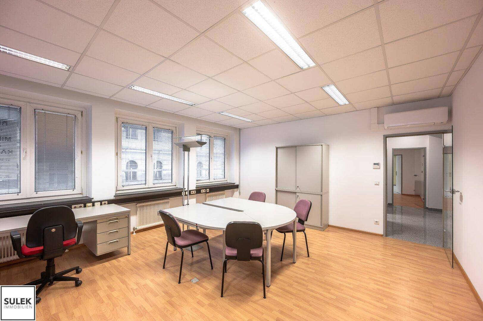 Helles Büro mit Besprechungstisch, Schränken und Blick auf die Stadt durch die Fenster.