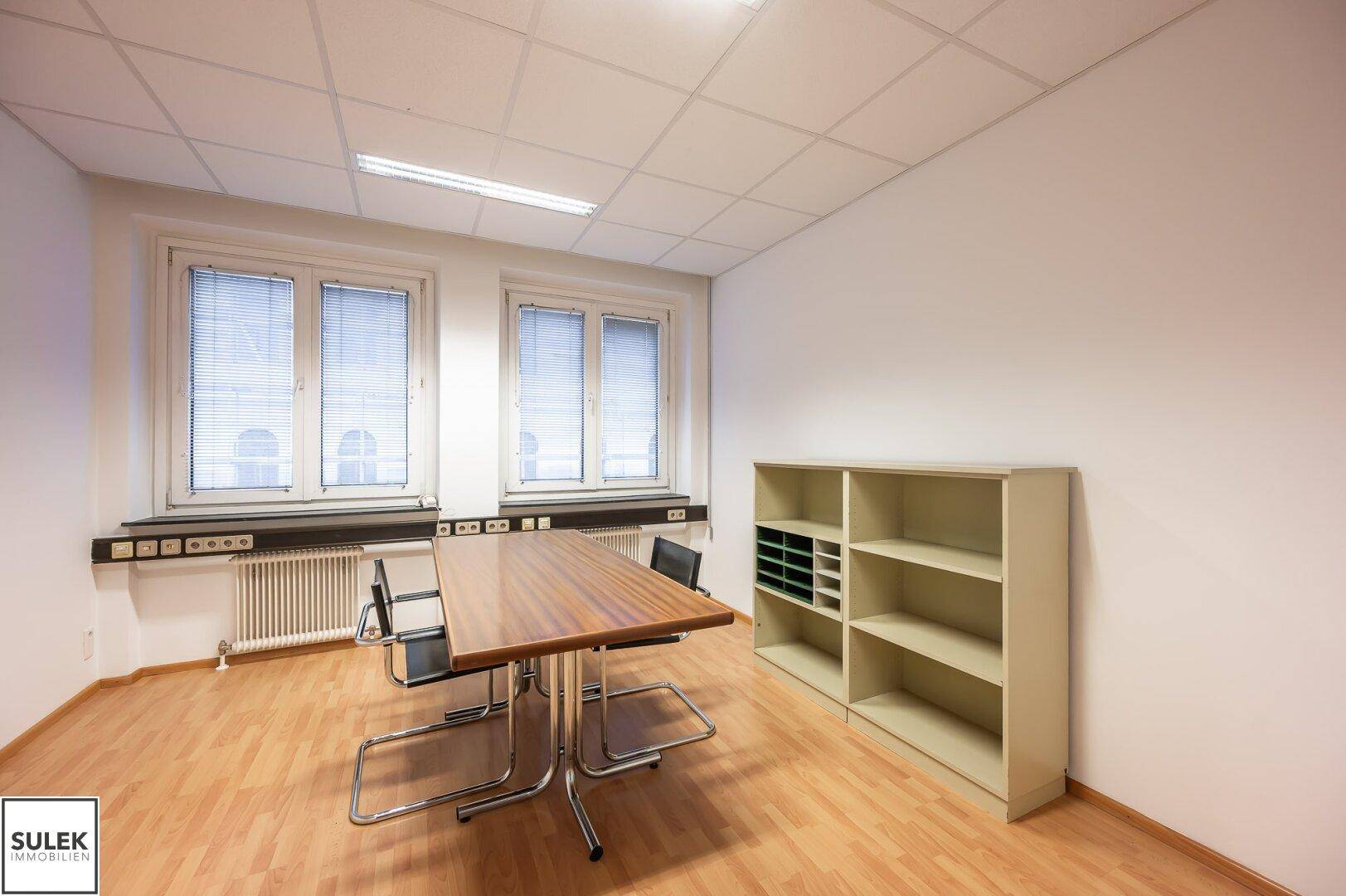Büro mit Holztisch, Regalen und großen Fenstern, die viel Tageslicht hereinlassen.