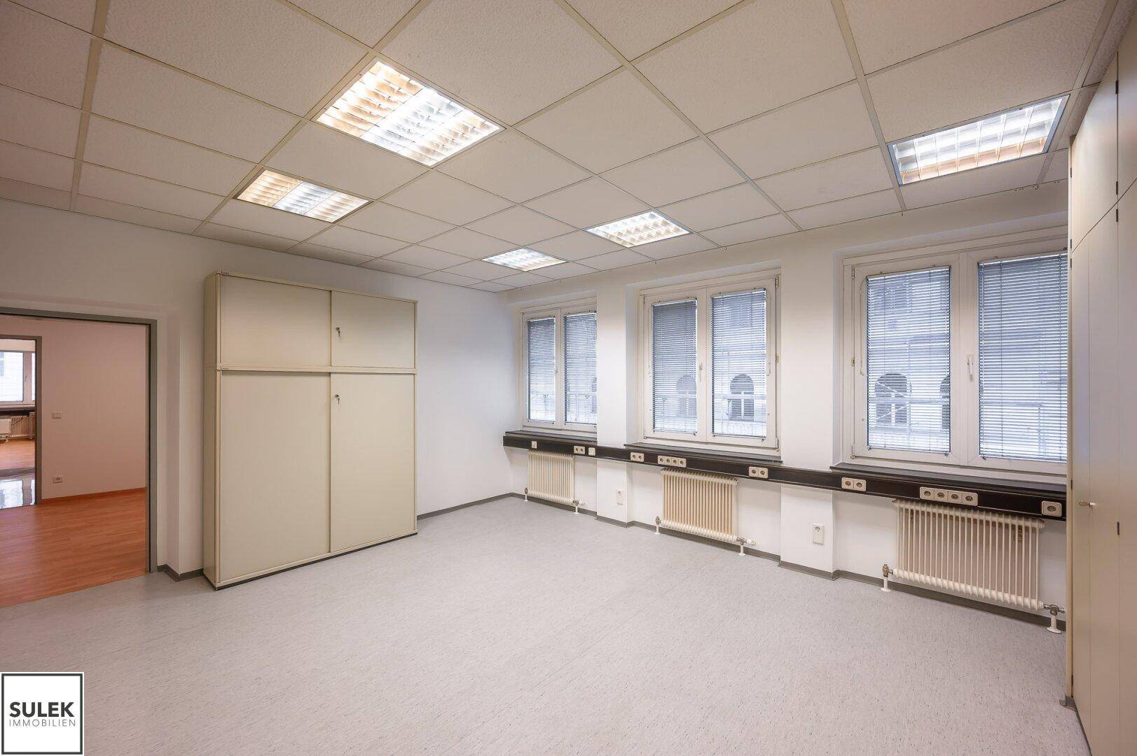 Leeres Büro mit Teppichboden, Einbauschränken und vielen Fenstern für natürliches Licht.