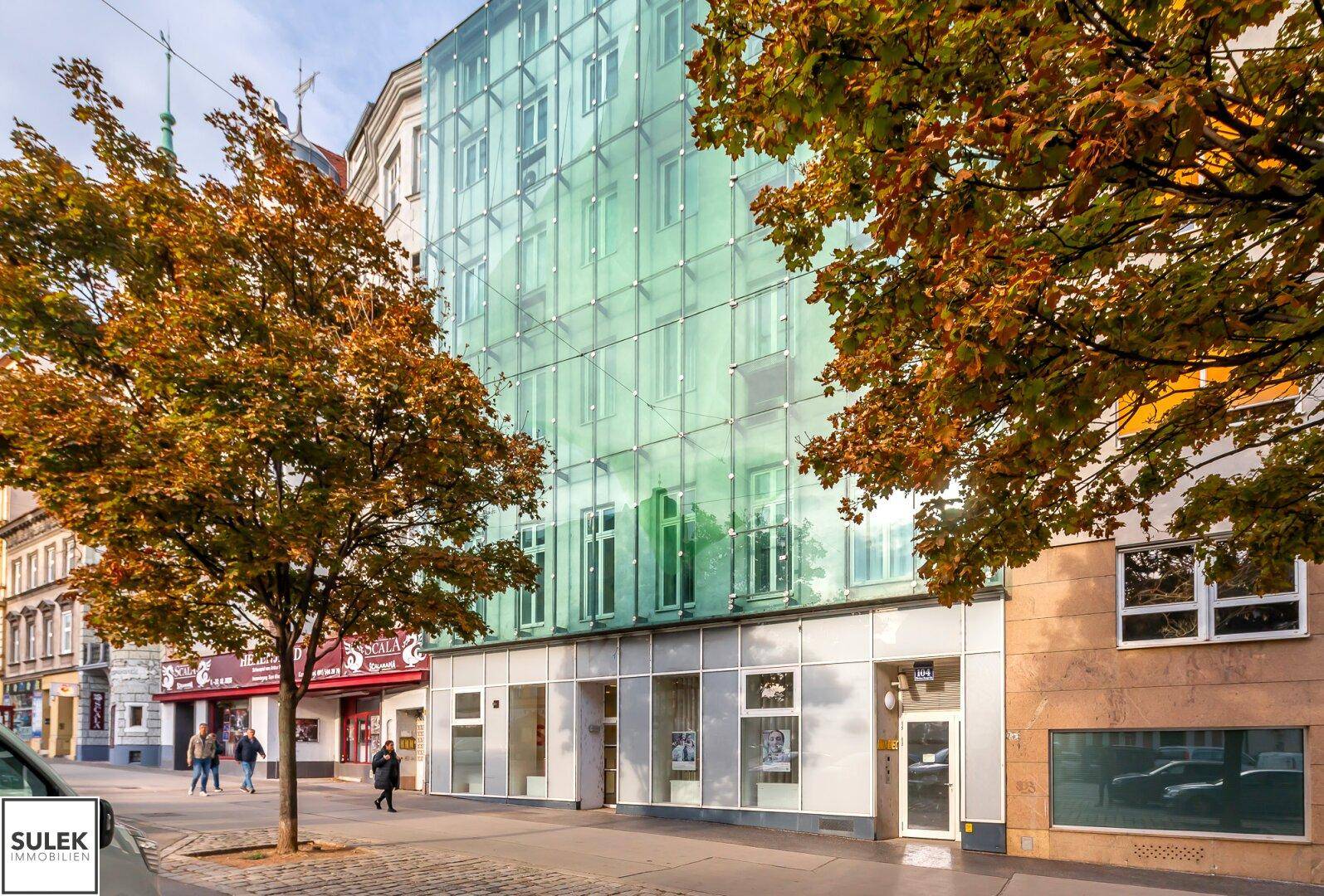 Moderne Glasfassade eines Geschäftsgebäudes mit Bäumen im Vordergrund, an einer belebten Straße.