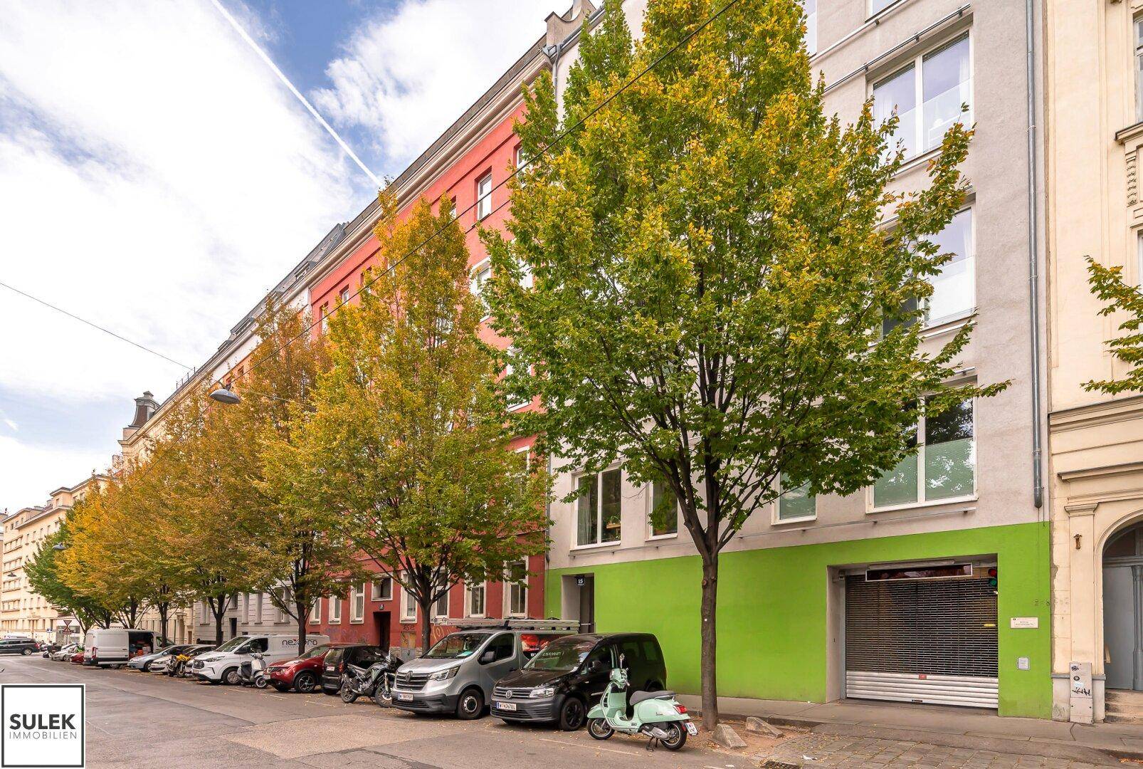 Straßenansicht eines Wohngebäudes mit Bäumen und parkenden Autos, grüne Fassadendetails.