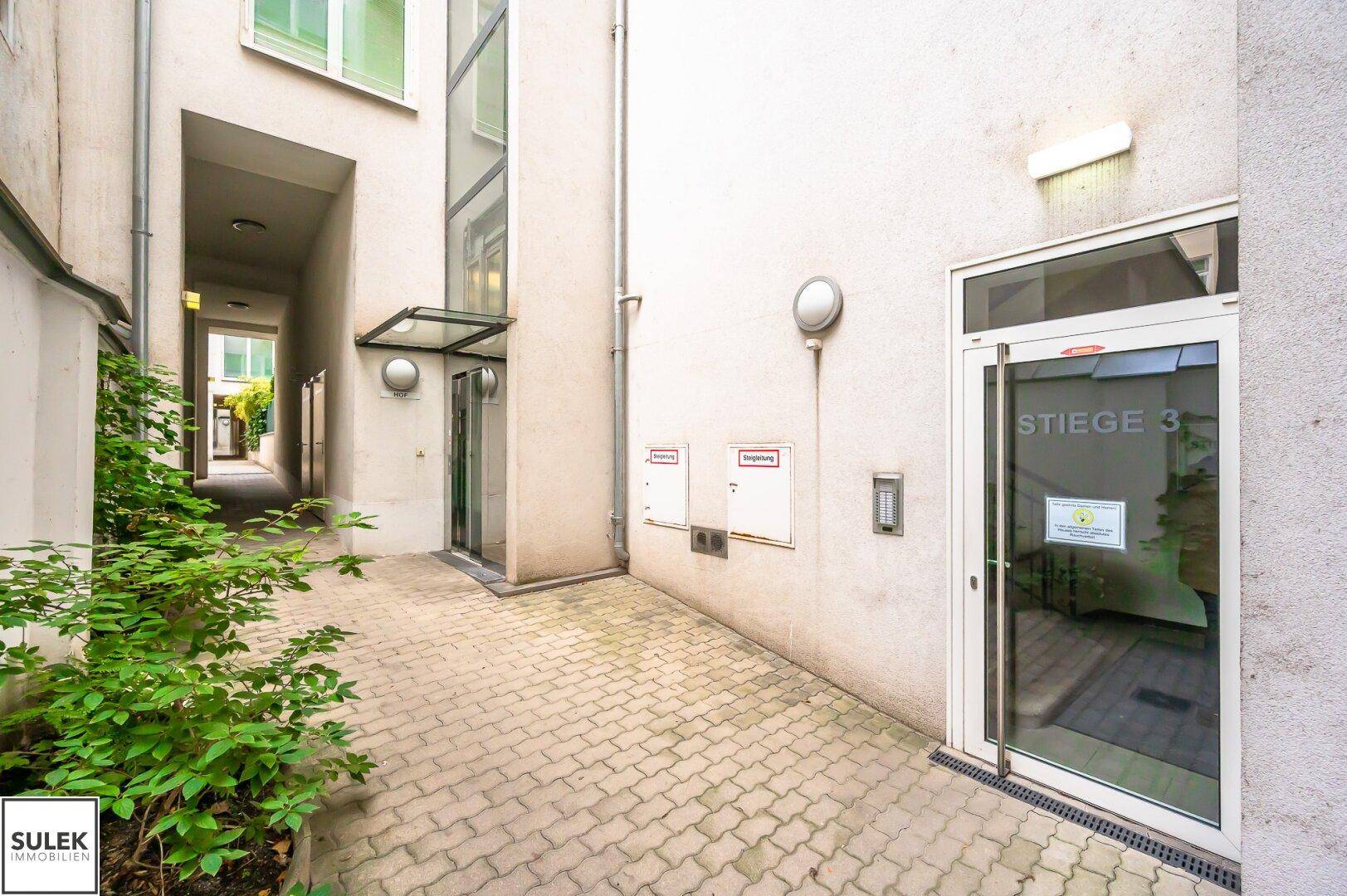 Gepflasterter Innenhof mit Pflanzen und Zugang zu den Gebäudeeingängen und einem Aufzug.
