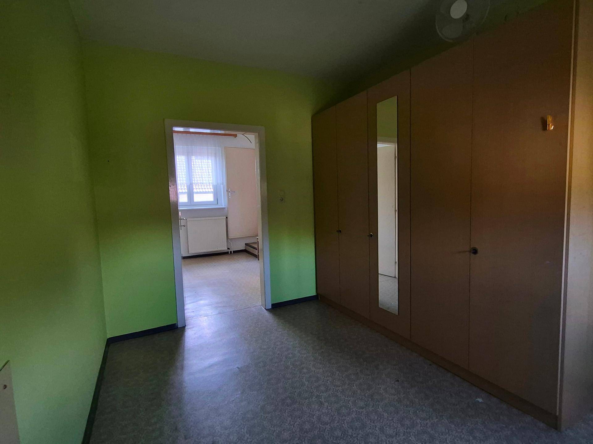 Einfaches Zimmer mit grünen Wänden, Schrank mit Spiegeltüren und Blick in einen weiteren Raum.