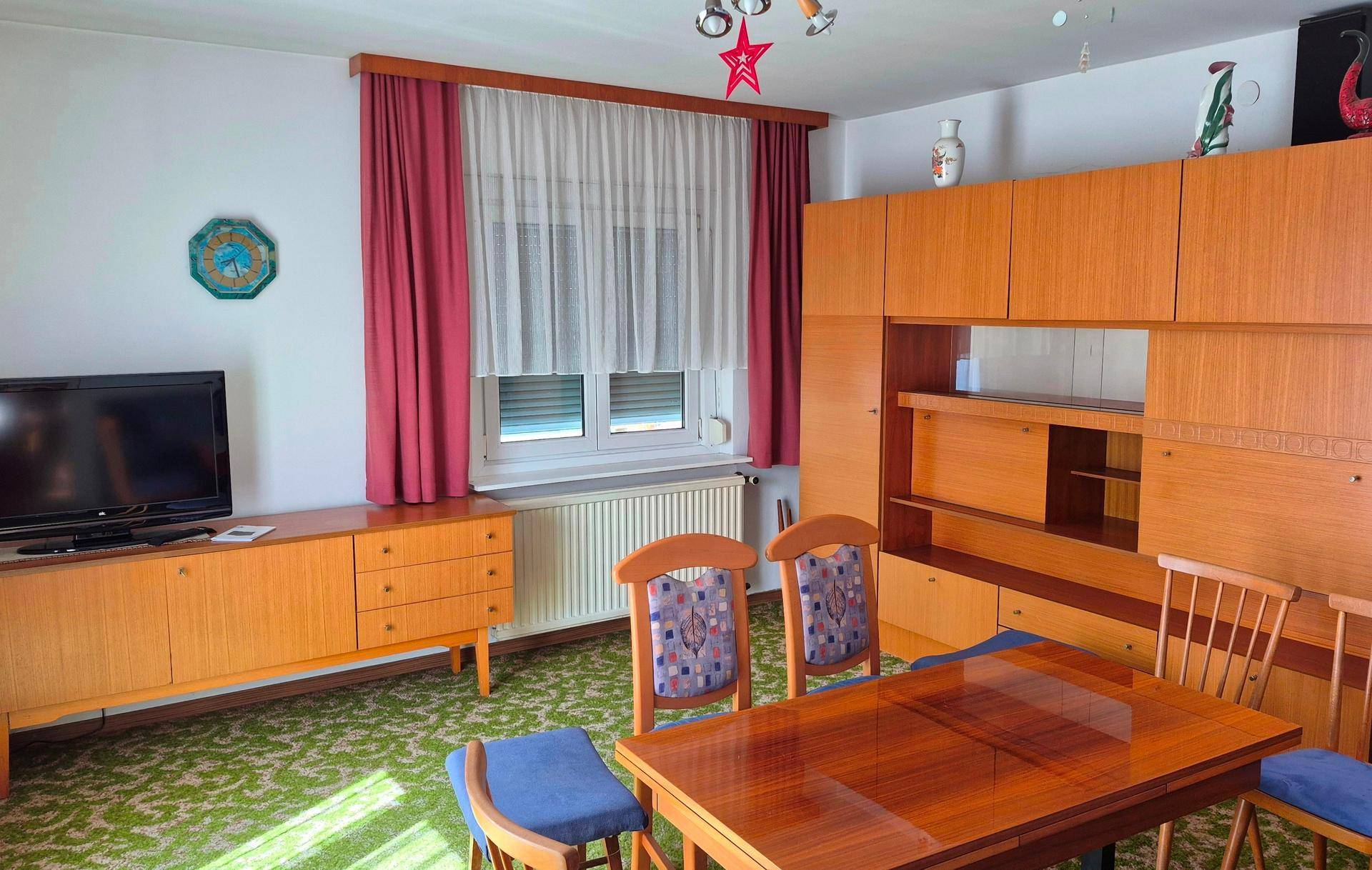 Gemütliches Wohnzimmer mit Teppichboden, Holzmöbeln und Fenster mit roten Vorhängen.