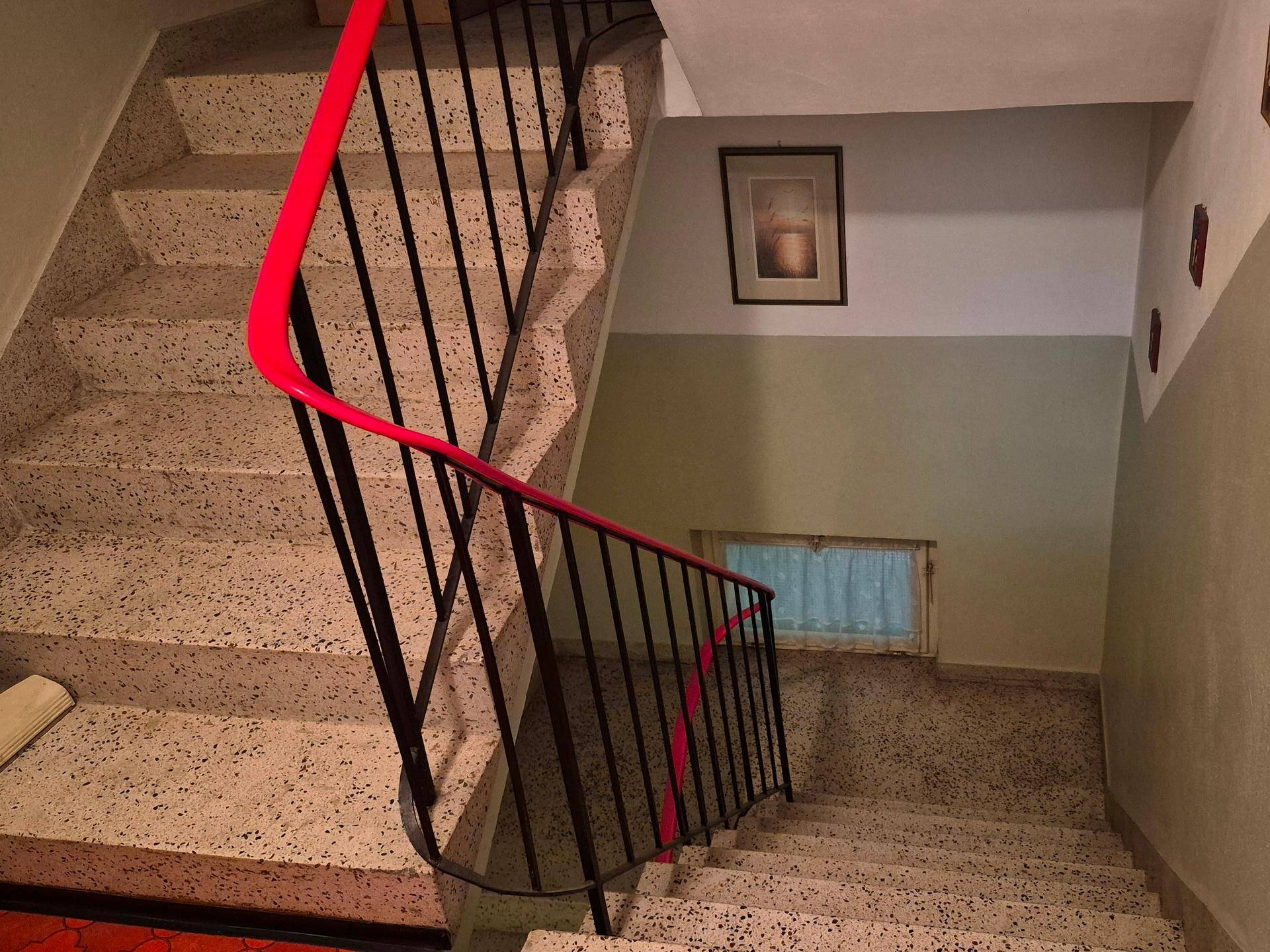 Treppenhaus mit Terrazzostufen und rotem Geländer, das die verschiedenen Etagen verbindet.