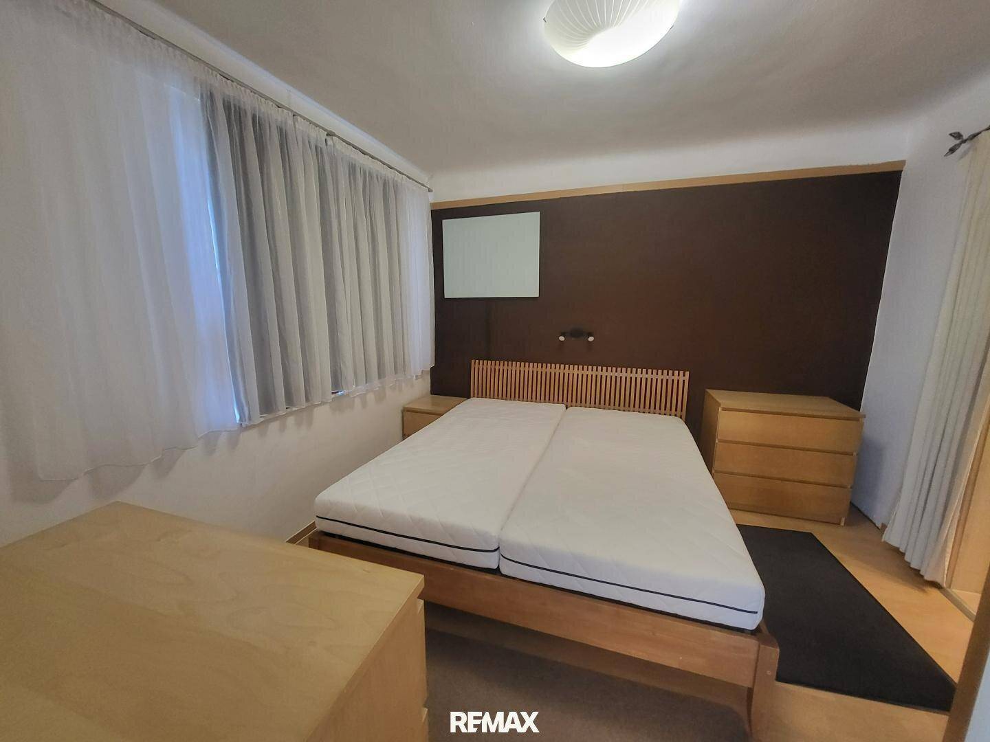 Schlafzimmer mit Doppelbett, Holzboden und Fenster mit weißen Vorhängen.