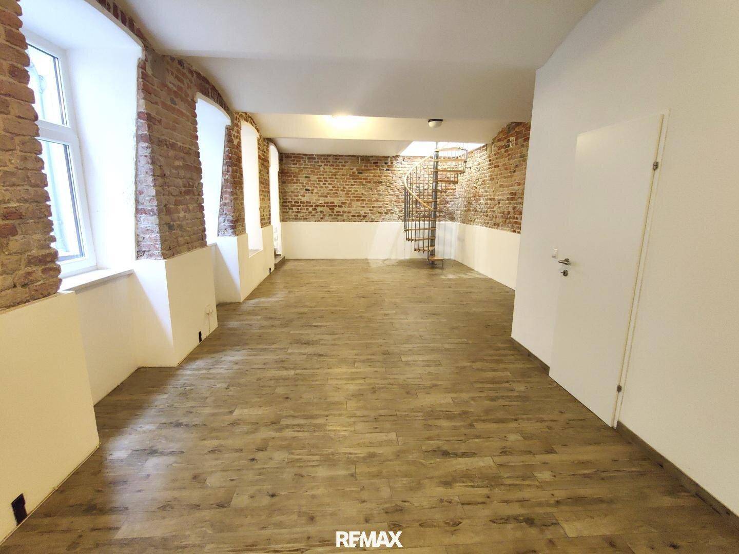 Großer Raum mit freiliegenden Ziegelwänden, Holzoptik-Boden und einer Wendeltreppe im Hintergrund.
