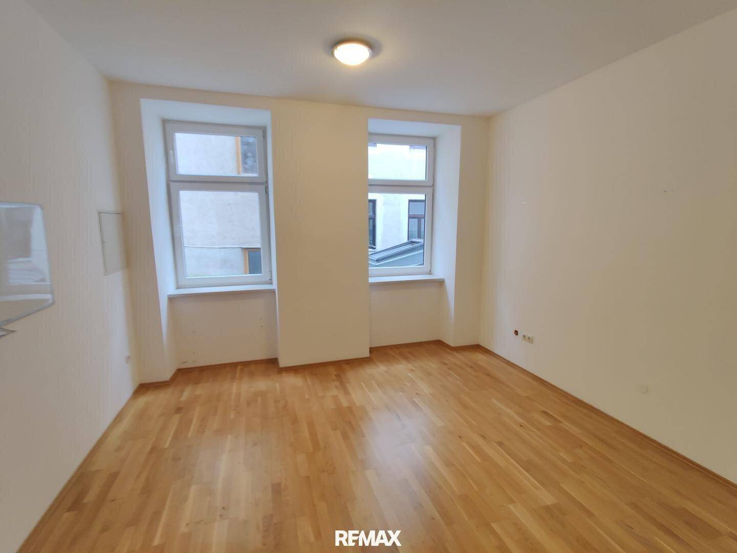 Leeres Zimmer mit hellem Holzparkettboden und zwei Fenstern, die viel Tageslicht hereinlassen.