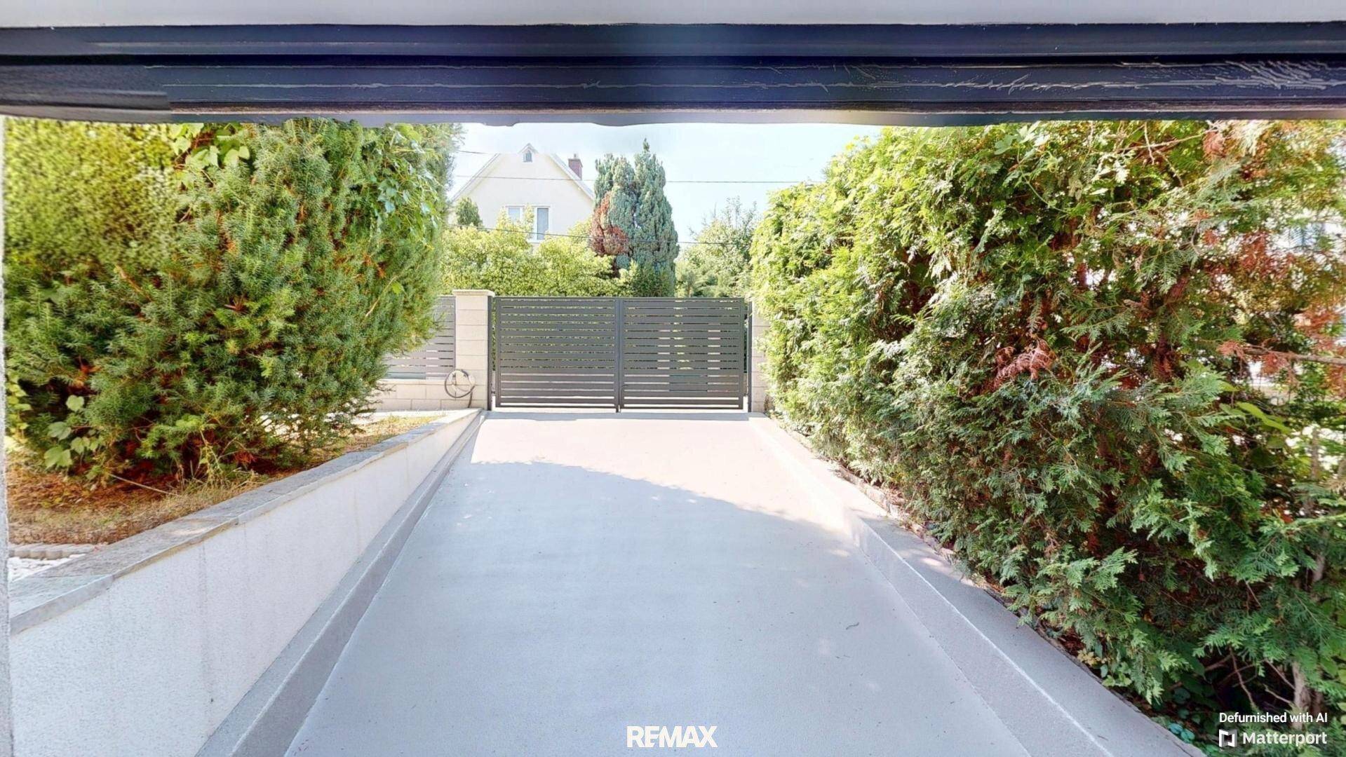 Blick von einem überdachten Bereich auf die Einfahrt mit modernem Tor und grünen Hecken.