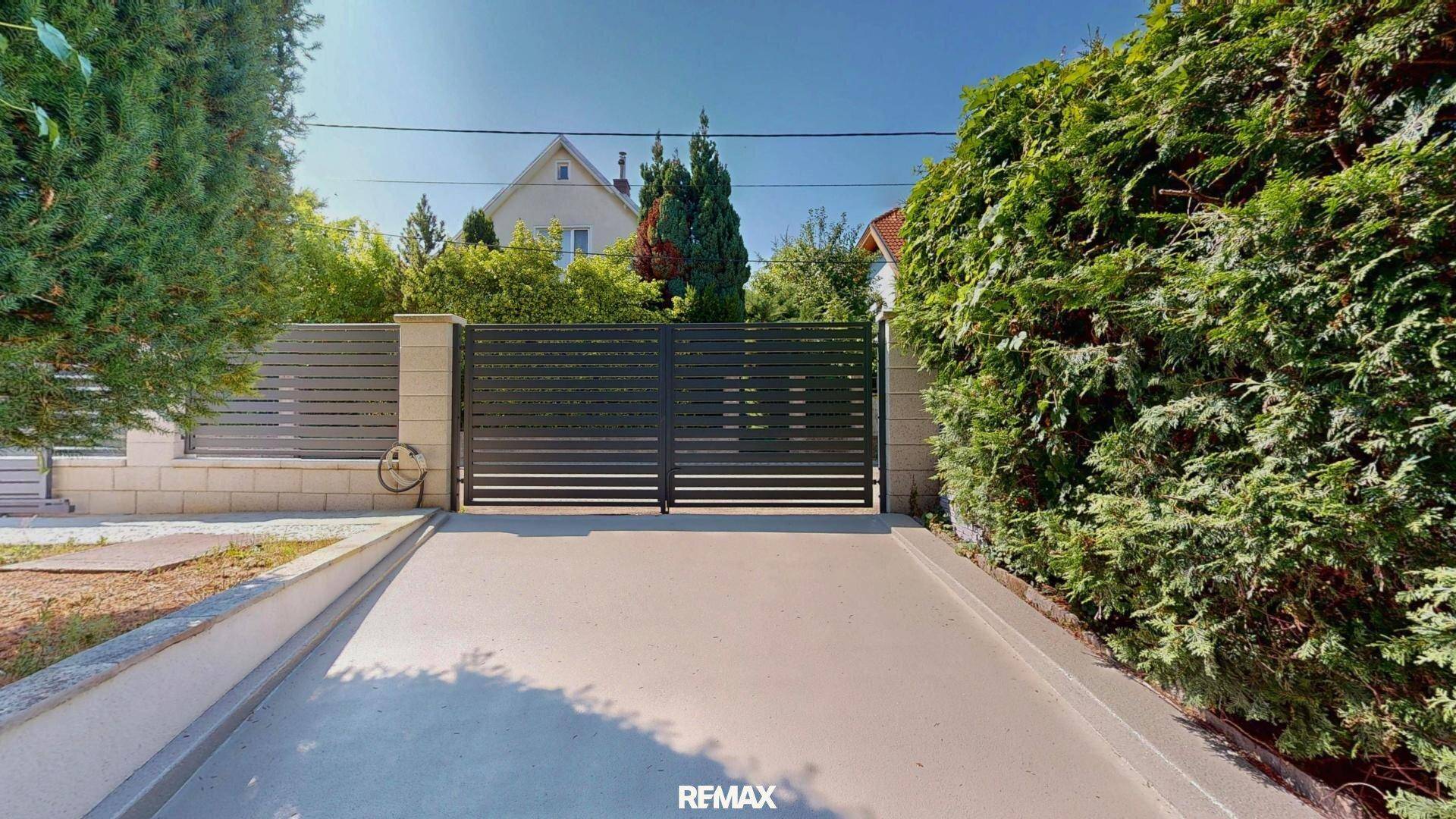 Breite Einfahrt mit einem modernen, dunklen Tor, umgeben von hohen, grünen Hecken.