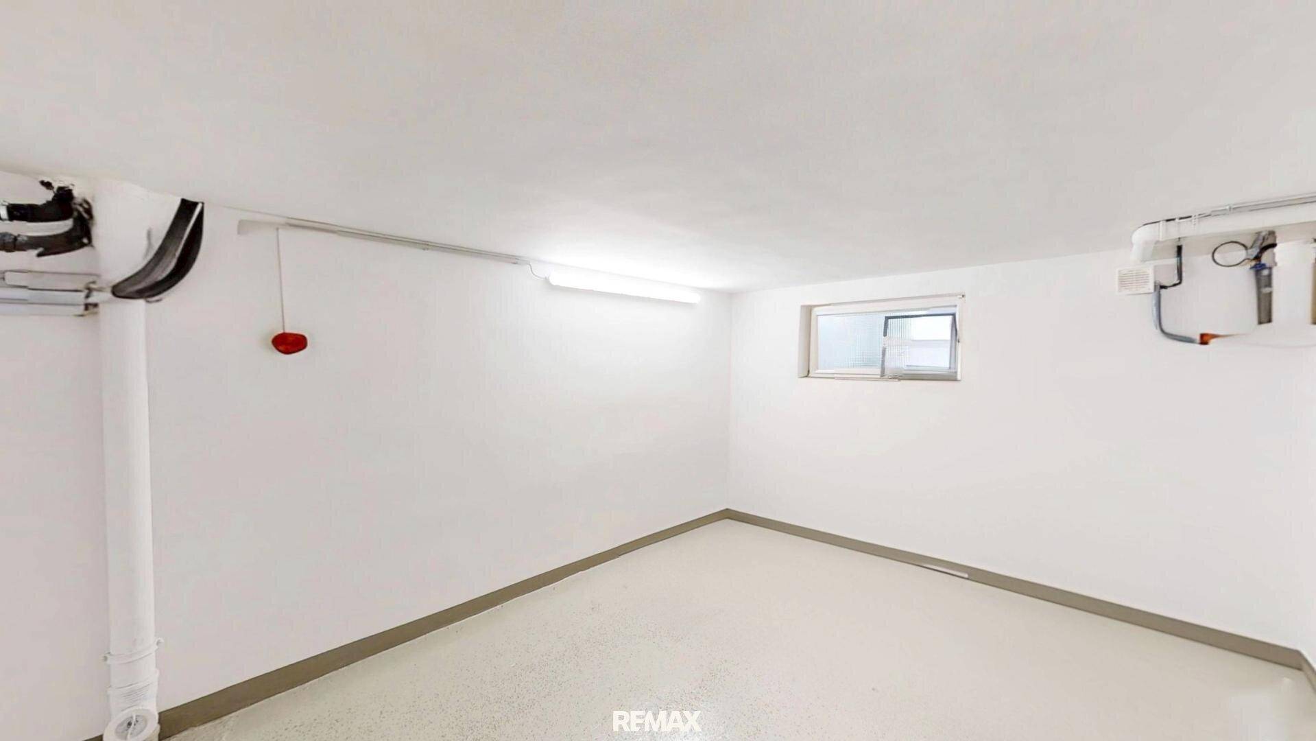 Heller, leerer Raum mit kleinem Fenster und sichtbaren Leitungen an der Decke.