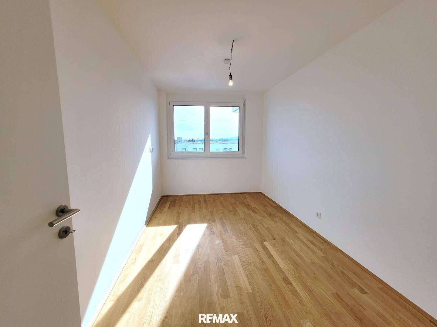 Helles Schlafzimmer mit Parkettboden und großem Fenster, das viel Tageslicht hereinlässt.