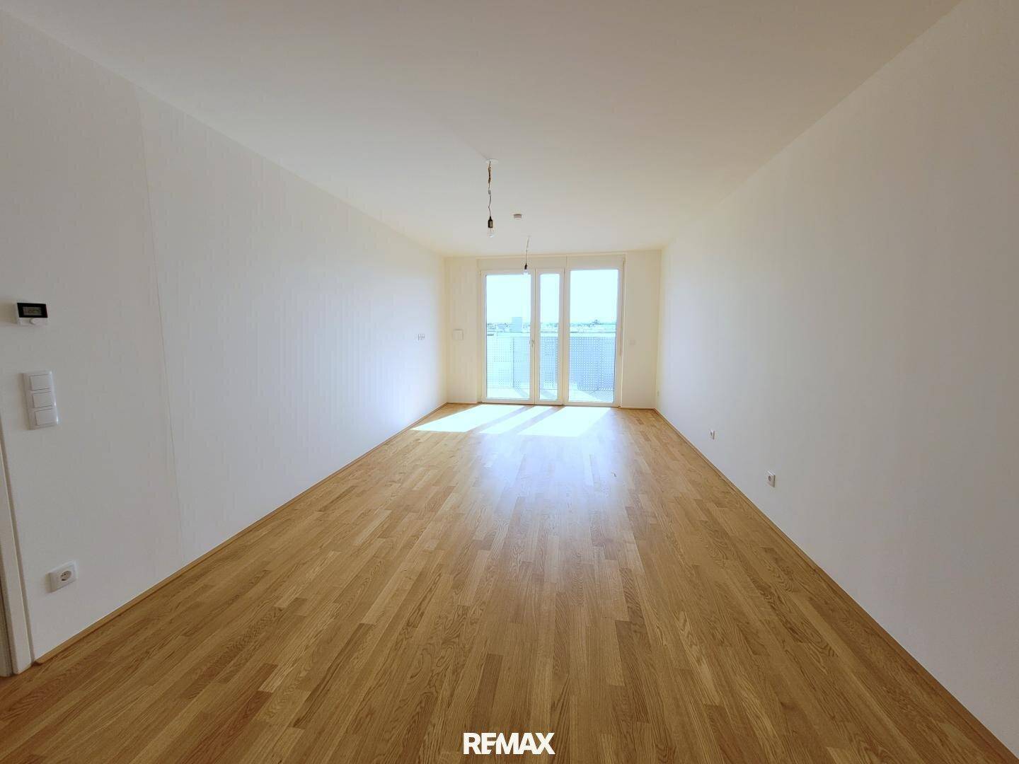 Lichtdurchfluteter Wohnraum mit hellem Parkettboden und Zugang zu einem Balkon.