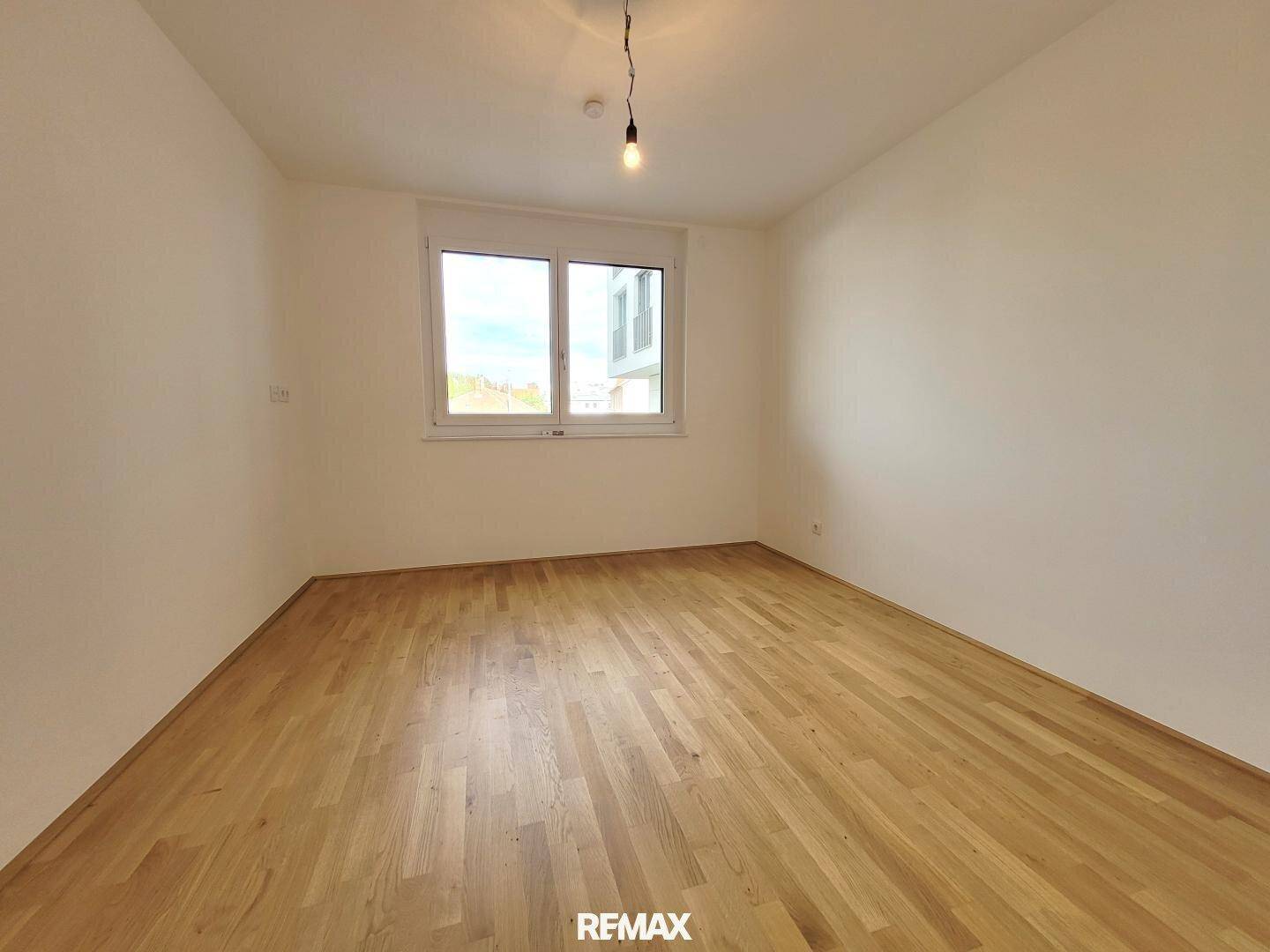 Leeres Schlafzimmer mit Parkettboden, weißen Wänden und einem großen Fenster für viel Tageslicht.
