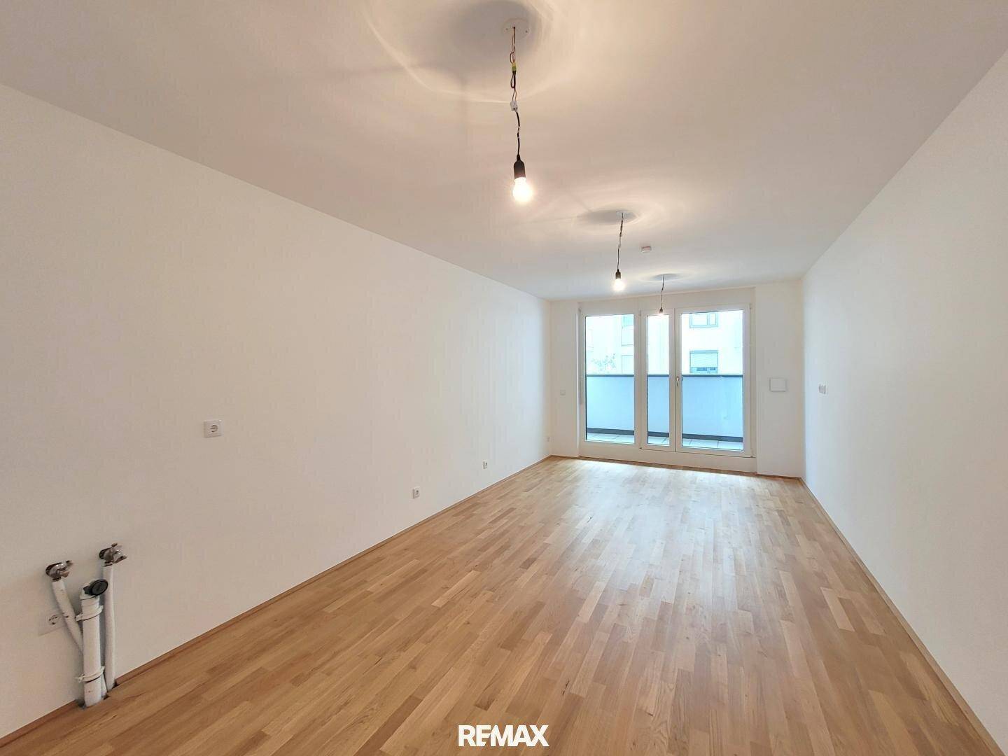 Geräumiger, leerer Raum mit Parkettboden und Zugang zu einem Balkon, vorbereitet für eine Küche.