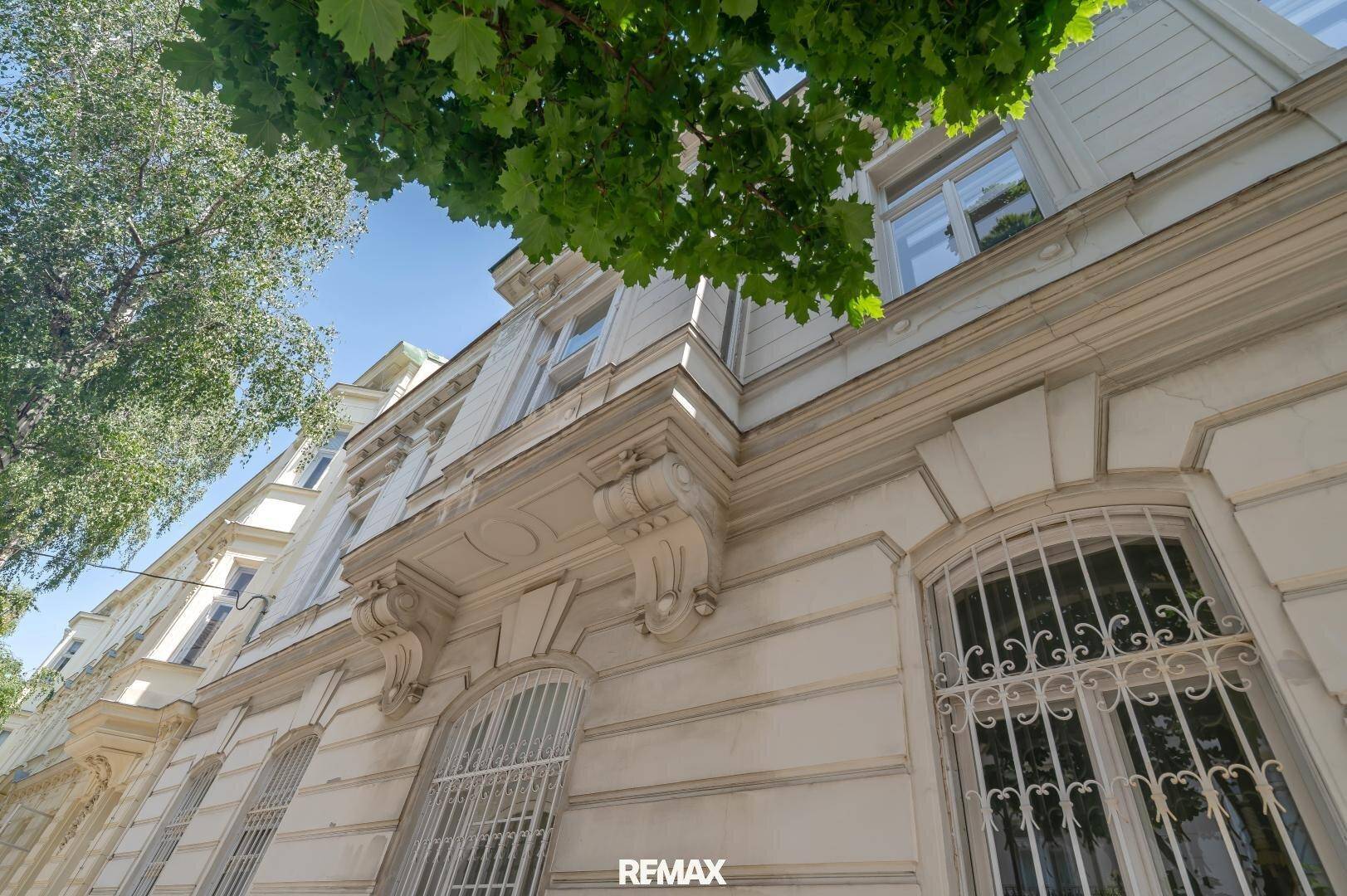 Elegante historische Fassade mit Stuckelementen, großen Fenstern und grünen Bäumen.