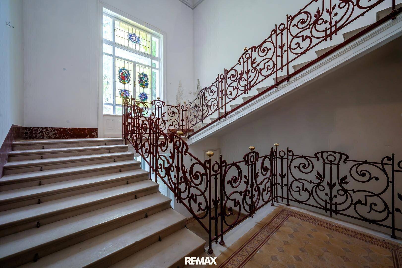 Elegantes Treppenhaus mit geschwungenem Geländer, Steinstufen und Buntglasfenster.