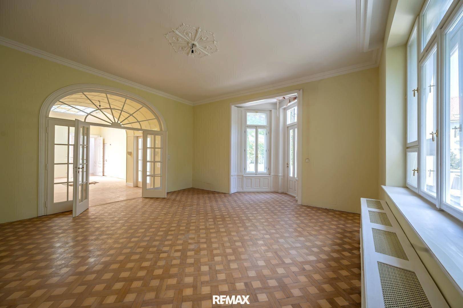 Geräumiges Wohnzimmer mit Parkettboden, Erkerfenster und Zugang zu einem weiteren Raum.