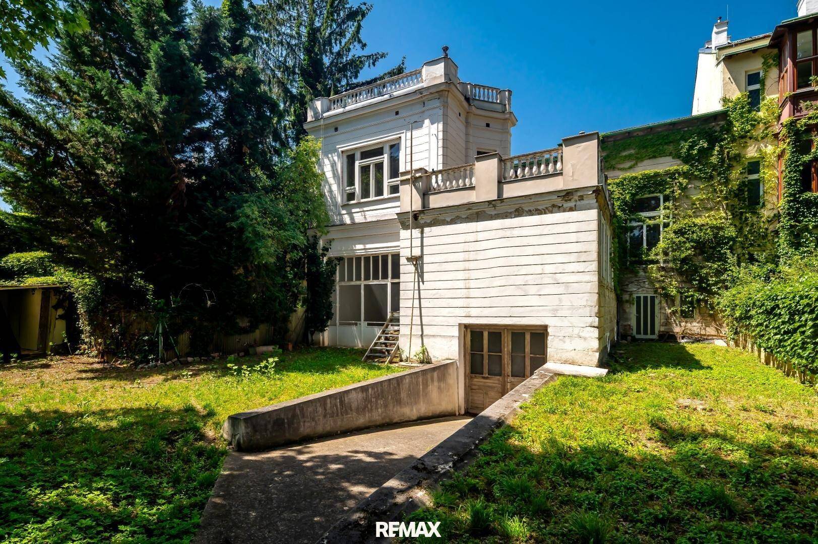 Rückansicht des historischen Gebäudes mit Garten, Kellerzugang und üppigem Grün.
