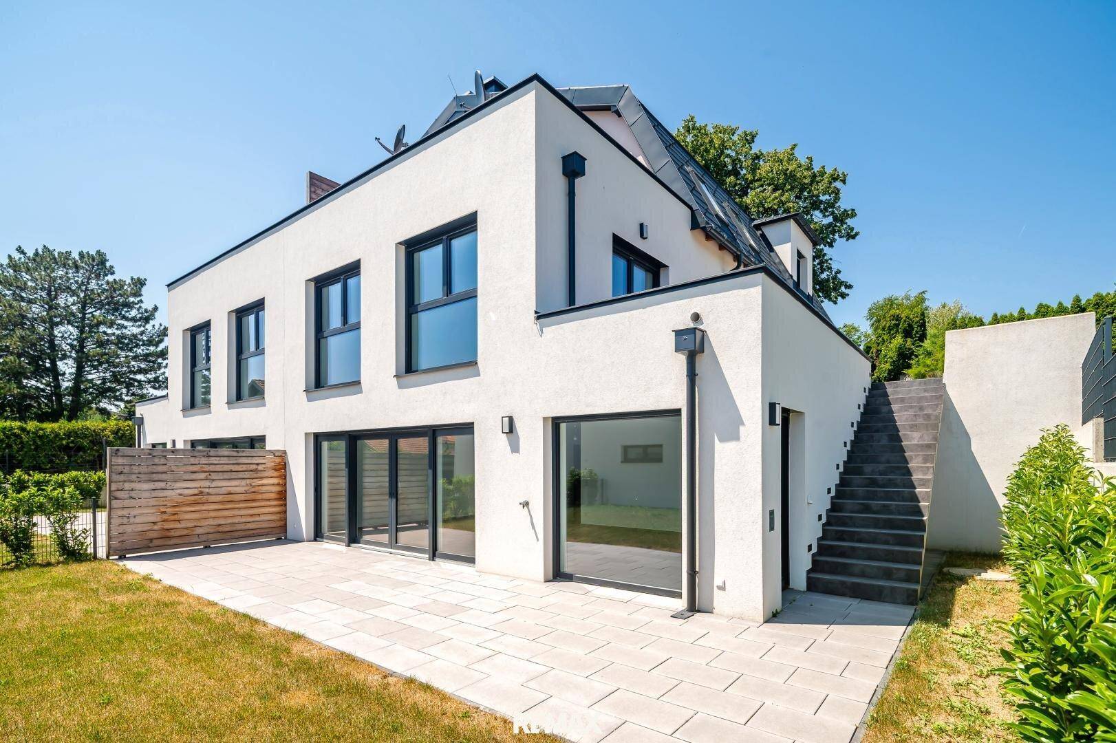 Moderne Hausfassade mit großer Terrasse und angrenzendem Gartenbereich unter blauem Himmel.