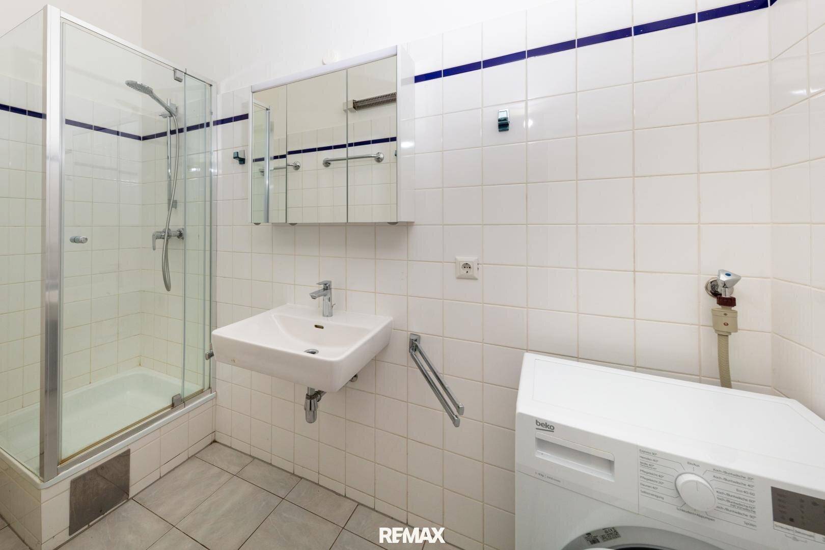 Funktionales Badezimmer mit weißen Fliesen, Duschkabine und Waschmaschine.