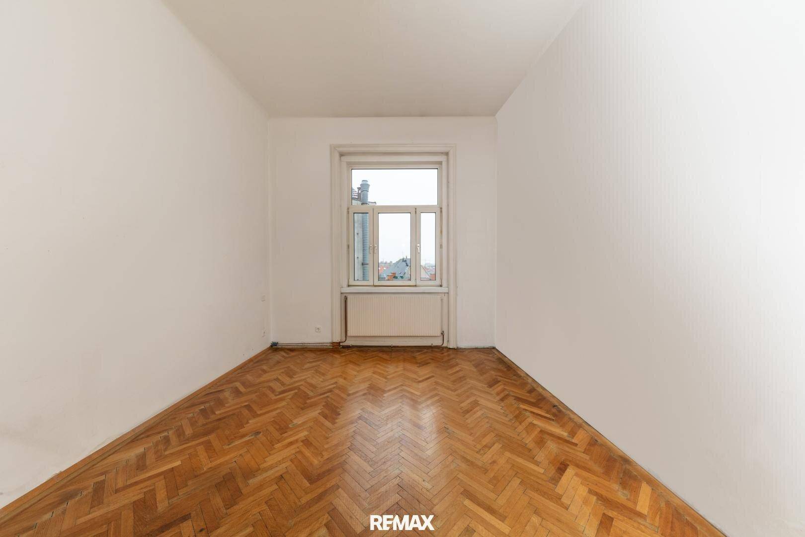 Schlichtes, leeres Zimmer mit Fischgrätparkett und einem Fenster, das auf die Stadt blickt.
