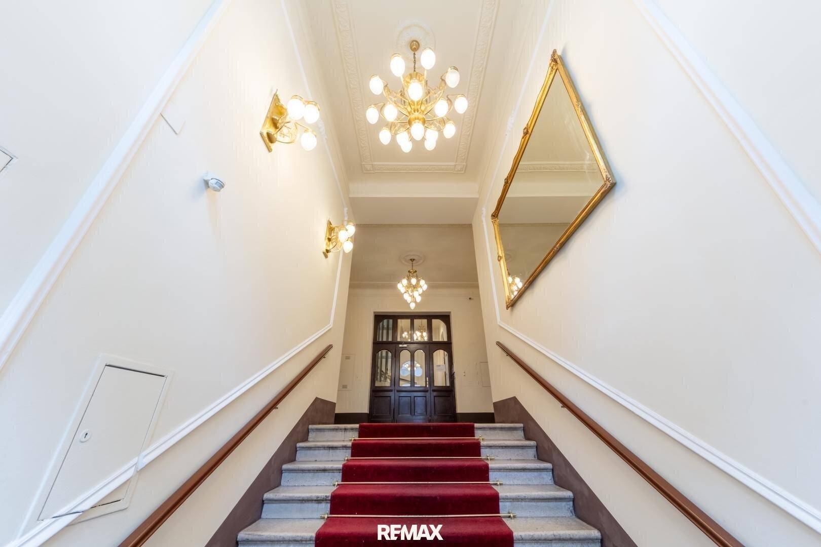 Elegantes Treppenhaus mit rotem Teppich, Kronleuchtern und Stuckdecke in einem Altbau.