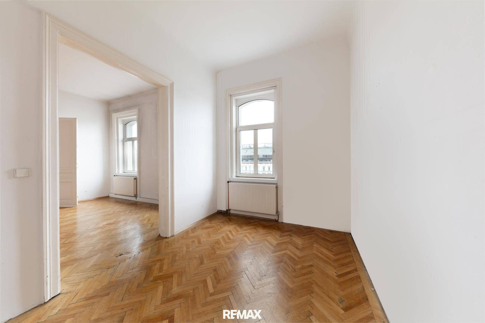 Leeres Zimmer mit Fischgrätparkett und hohen Fenstern, das viel Tageslicht hereinlässt.