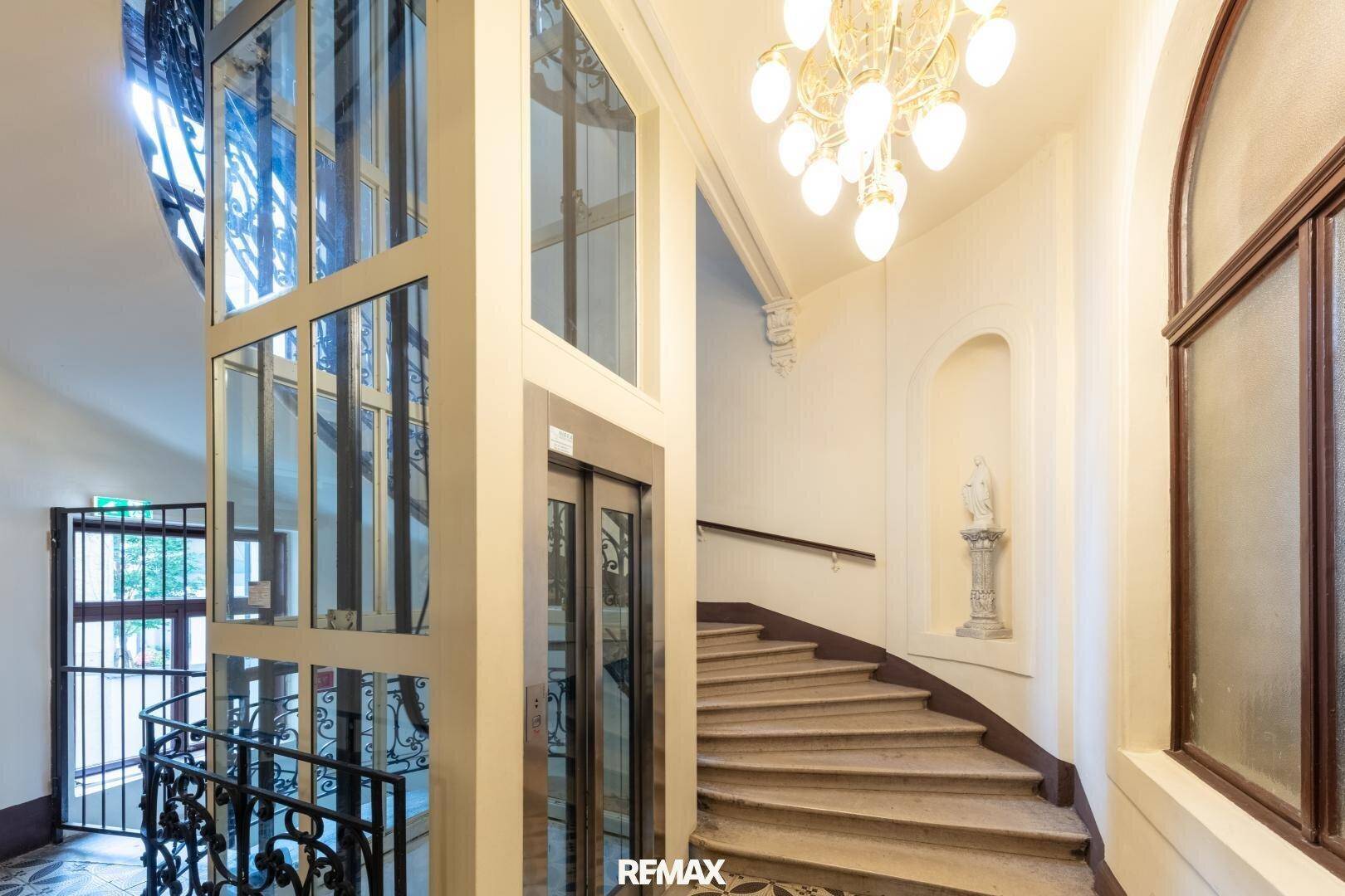 Modernisiertes Treppenhaus mit Glasaufzug und elegantem Kronleuchter in einem Altbau.