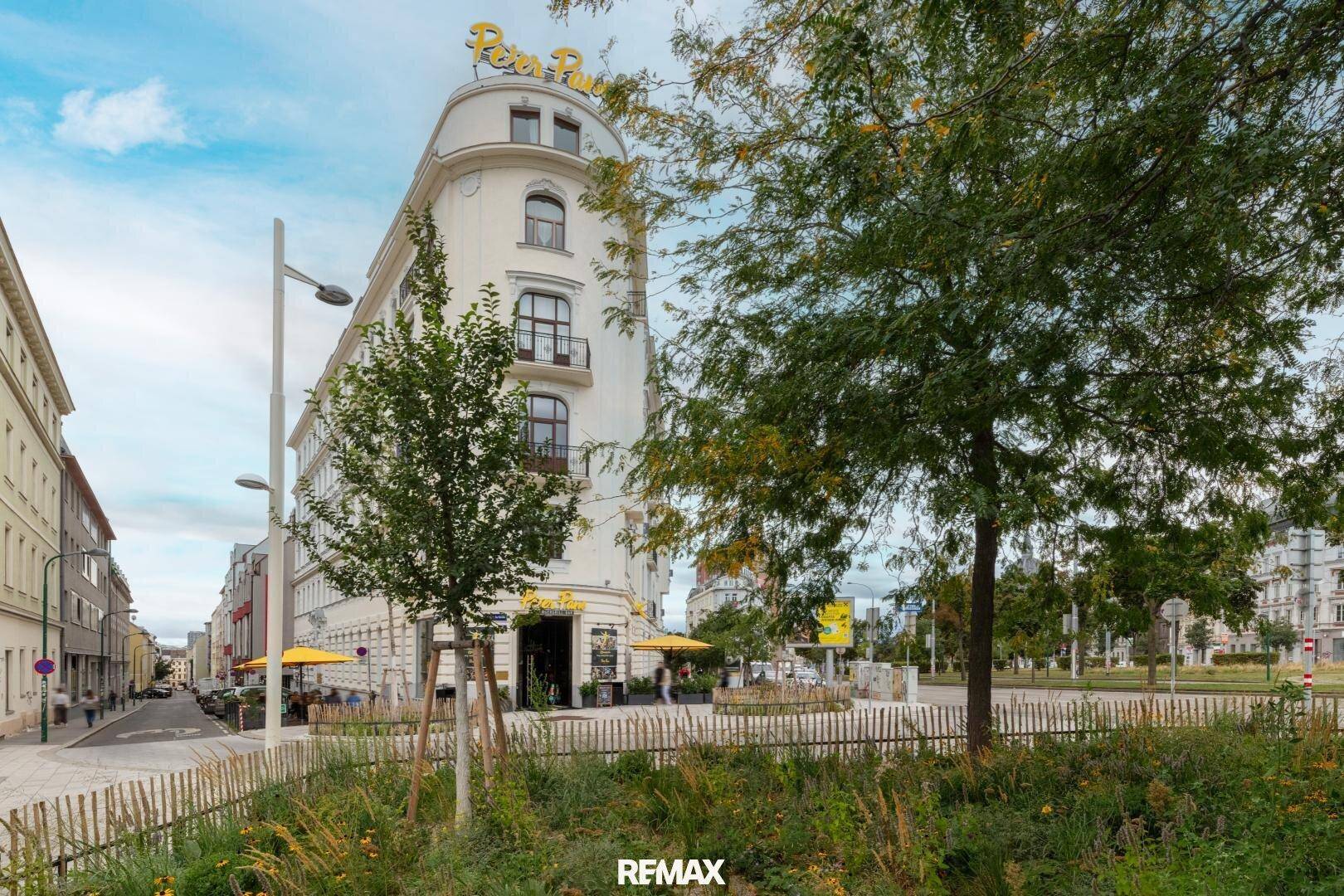 Historisches Eckgebäude mit Geschäften im Erdgeschoss und Bäumen im Vordergrund.