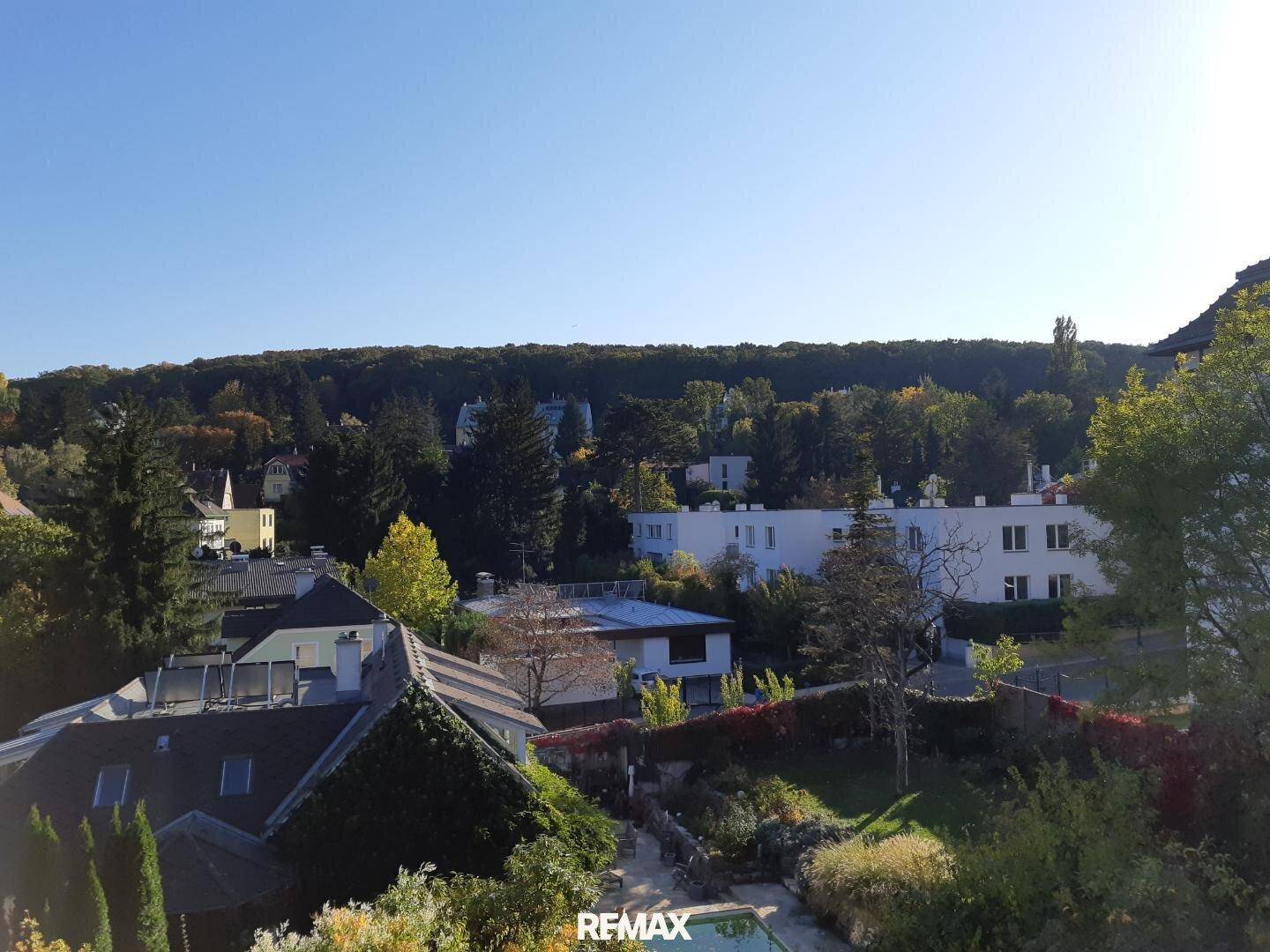 Panoramablick über eine Wohnsiedlung mit viel Grün und einem Wald im Hintergrund unter blauem Himmel.