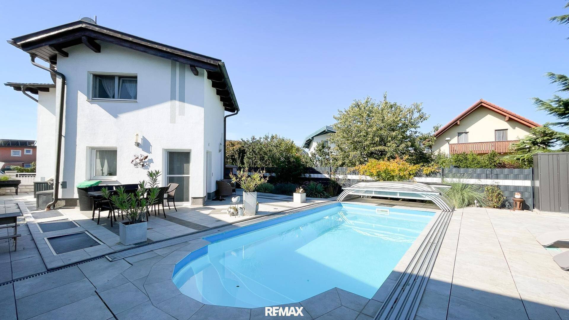Weißes Einfamilienhaus mit großem Poolbereich und gepflasterter Terrasse unter blauem Himmel.