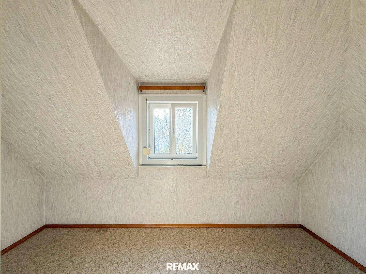 Dachzimmer mit schrägen Wänden, Teppichboden und einem Fenster, das den Raum erhellt.
