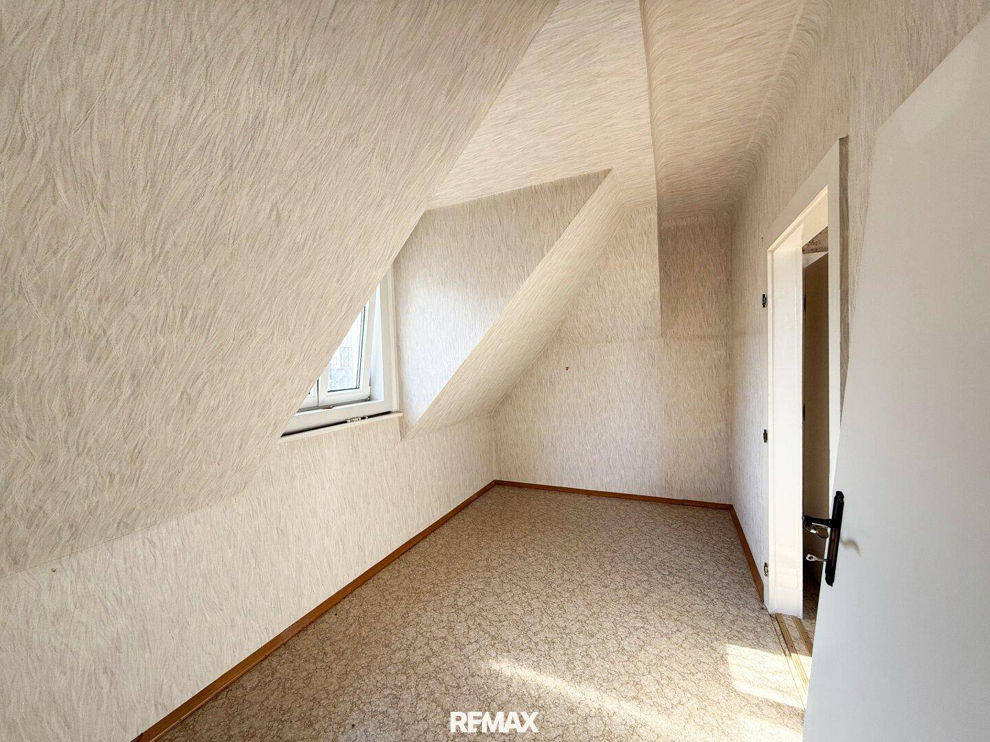 Helles Dachzimmer mit Teppichboden, schrägen Wänden und Fenster, das viel Tageslicht hereinlässt.