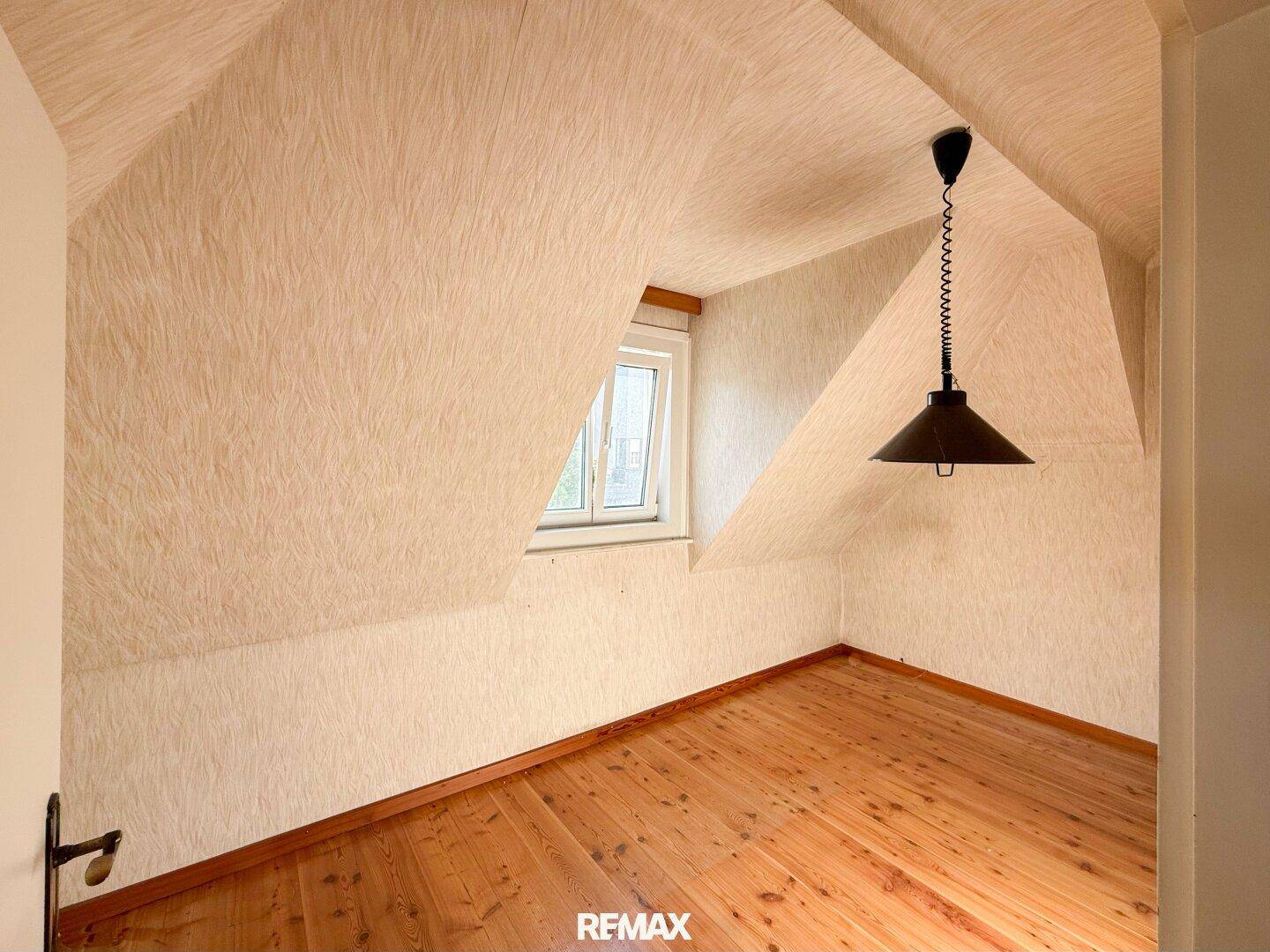 Dachzimmer mit Holzfußboden, schrägen Wänden und einem Fenster, das viel Tageslicht hereinlässt.