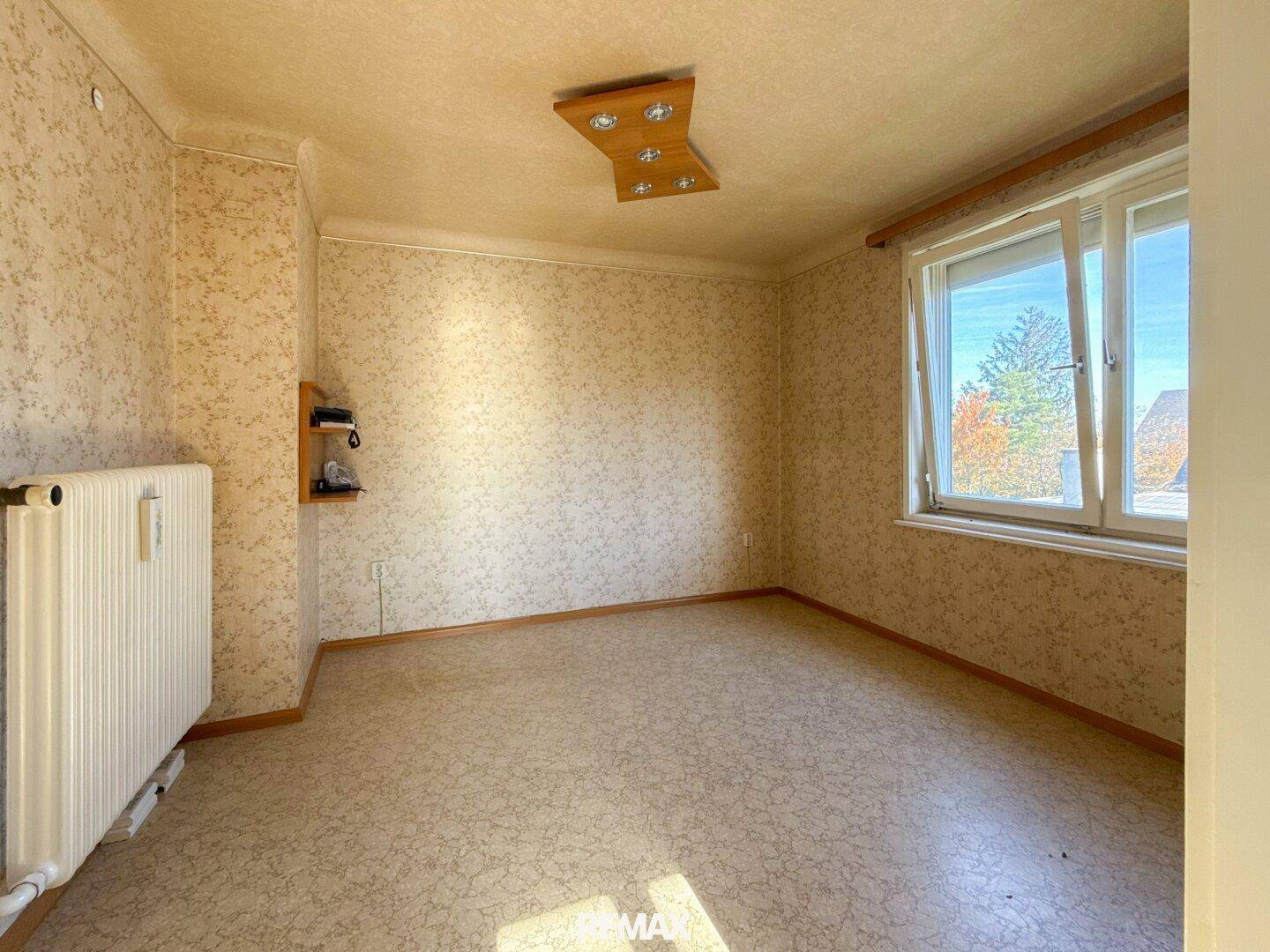 Helles Zimmer mit gemusterter Tapete, Korkboden und großem Fenster, das viel Licht spendet.