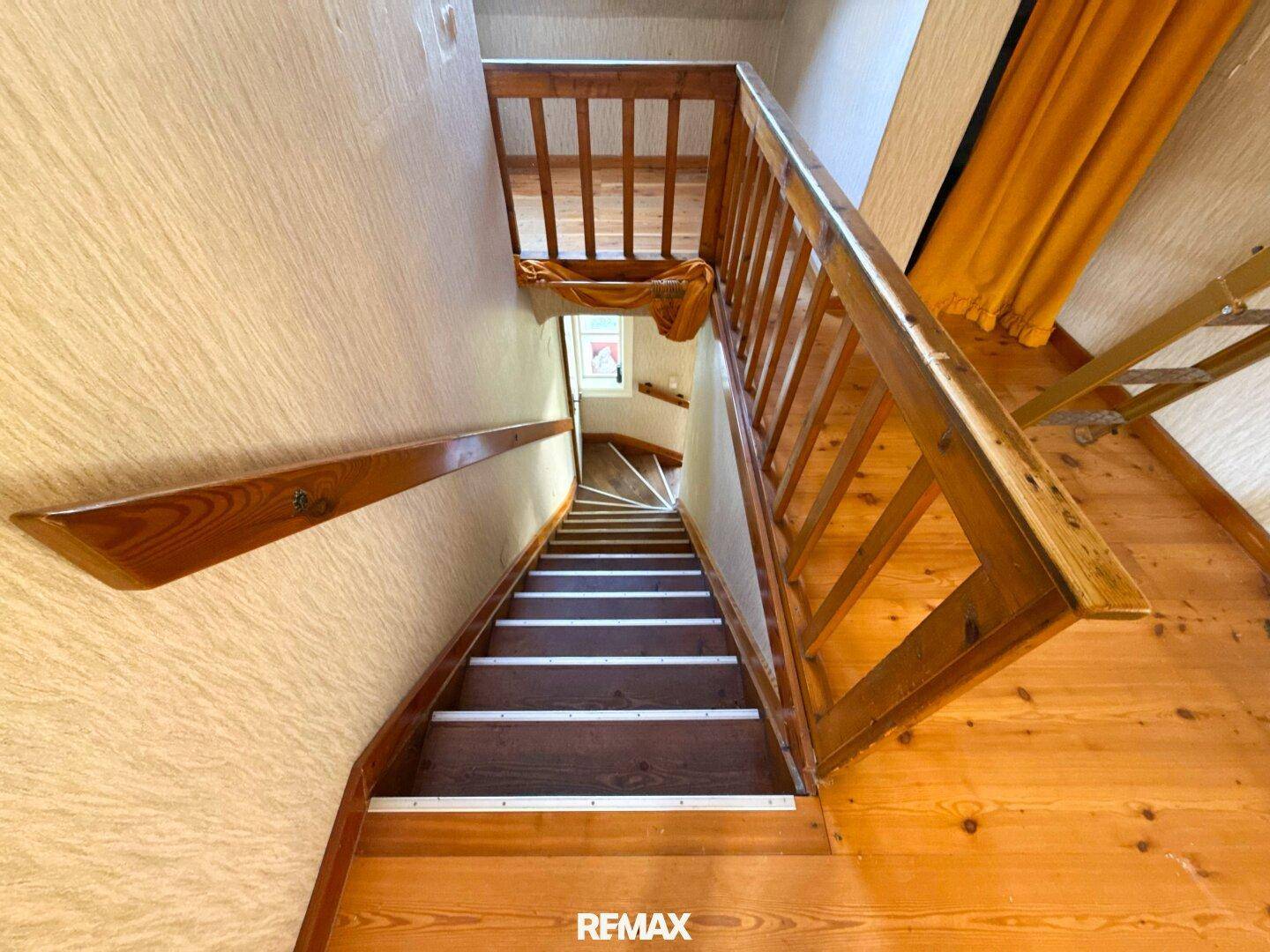 Holztreppe führt nach unten, mit Holzgeländer und Dachfenster im Hintergrund, das Licht spendet.