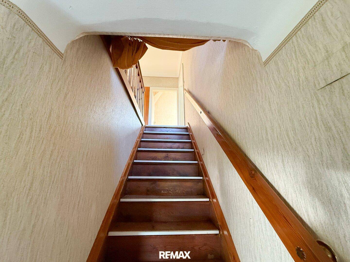Holztreppe führt nach oben in einen hellen Flur mit Tapeten, der zu weiteren Zimmern führt.