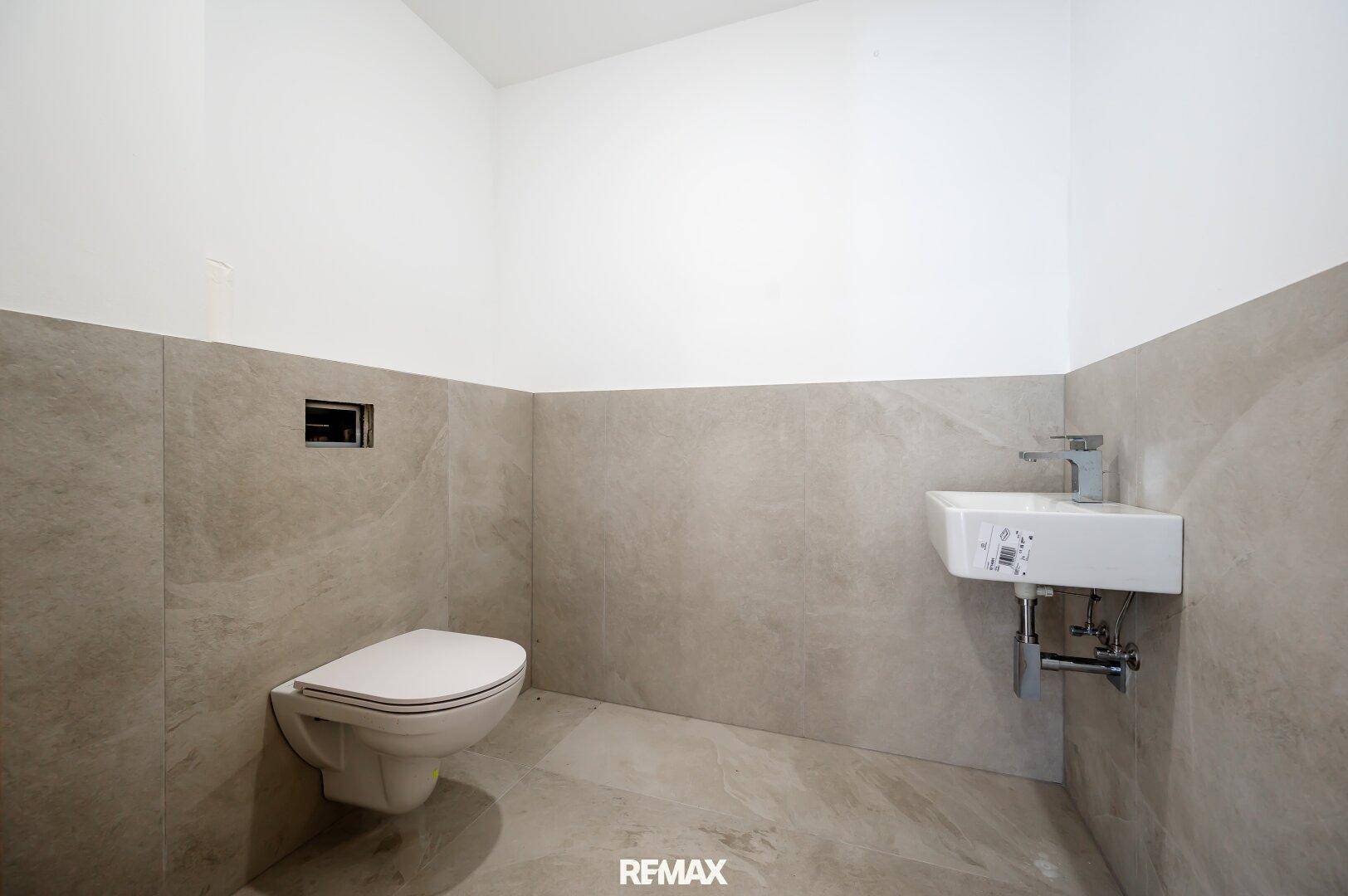 Modernes Gäste-WC mit Wand-WC und kleinem Waschbecken, halbhoch gefliest in neutralen Farben.