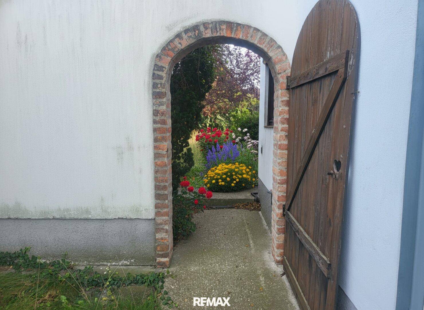 Rustikaler Ziegelbogen führt zu einem farbenfrohen Garten mit blühenden Blumen.