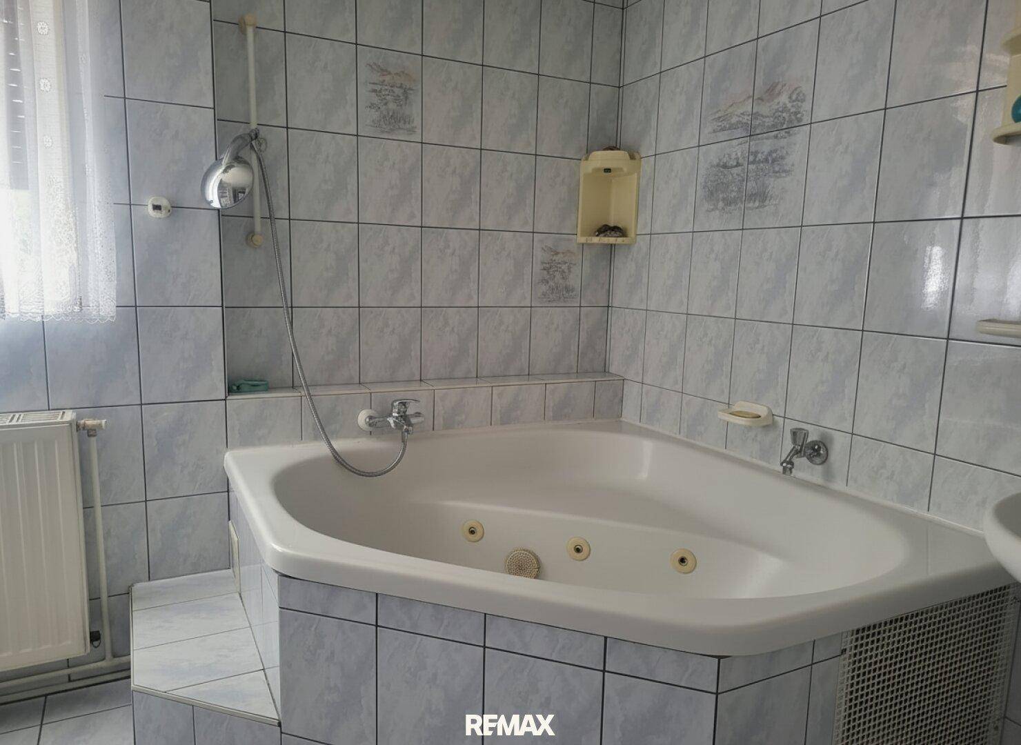 Gefliestes Badezimmer mit Eckbadewanne, Duschvorrichtung und Fenster.