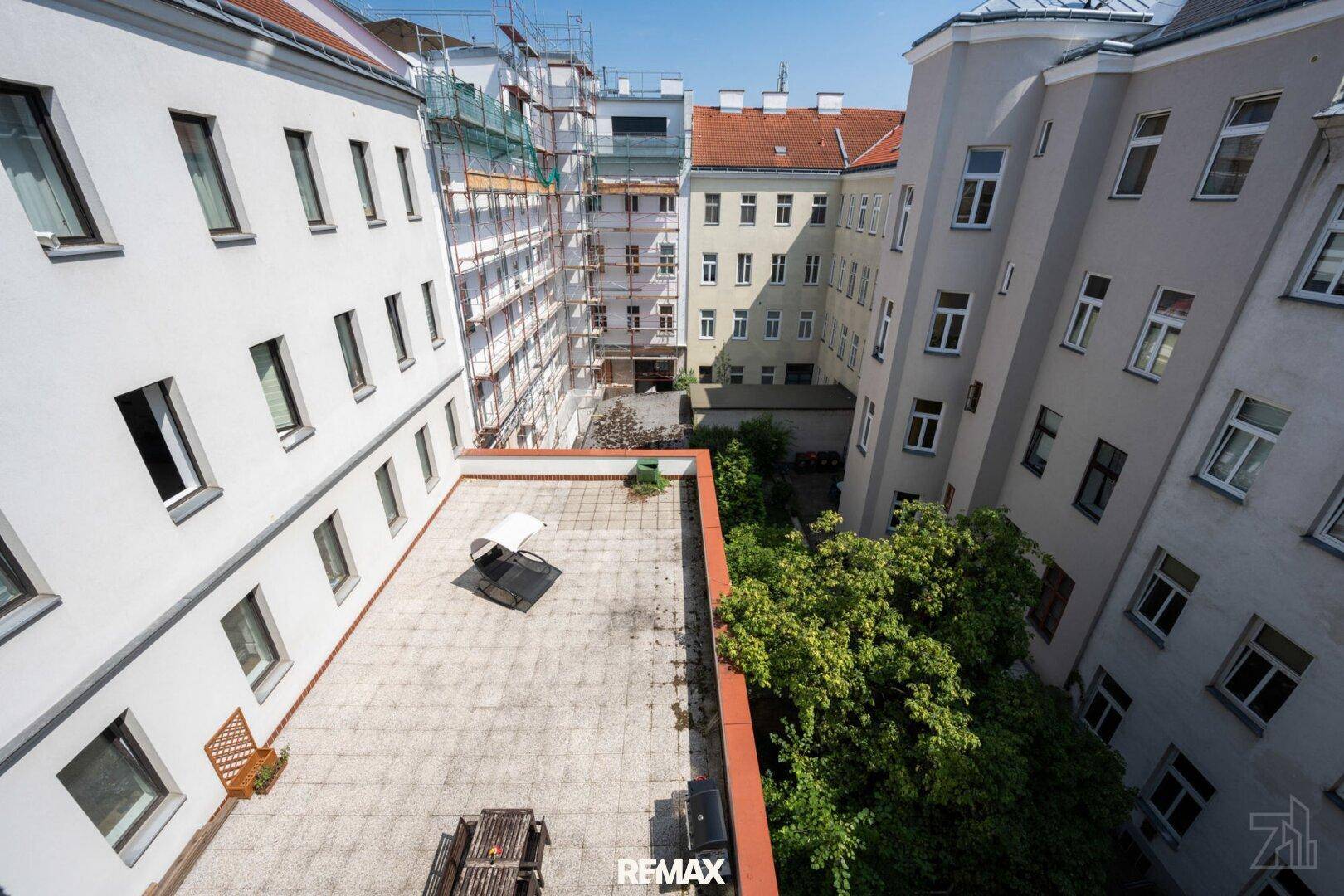 Große gepflasterte Terrasse im Innenhof mit Sitzgelegenheiten, umgeben von Gebäuden und Bäumen.